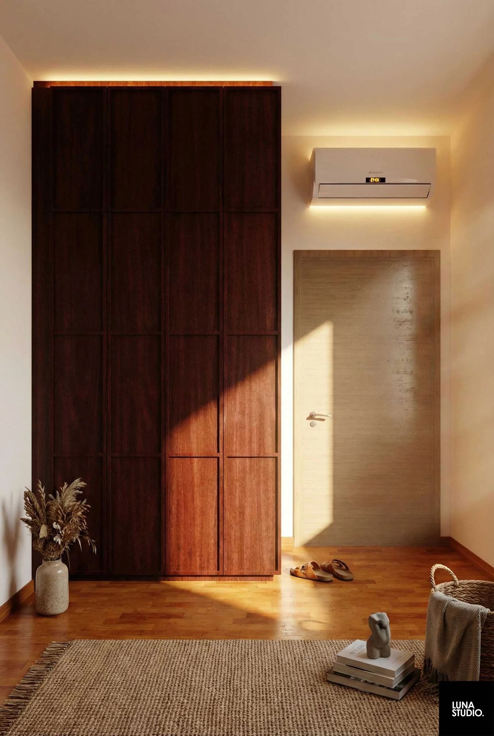 Interior of a room with wooden floor, a large dark wooden wardrobe, a beige door, a pair of sandals on the floor, a vase with dried flowers, a stack of books with a small sculpture on top, and a woven rug. An air conditioning unit is mounted on the wall above the door, with warm lighting and sunlight casting shadows.