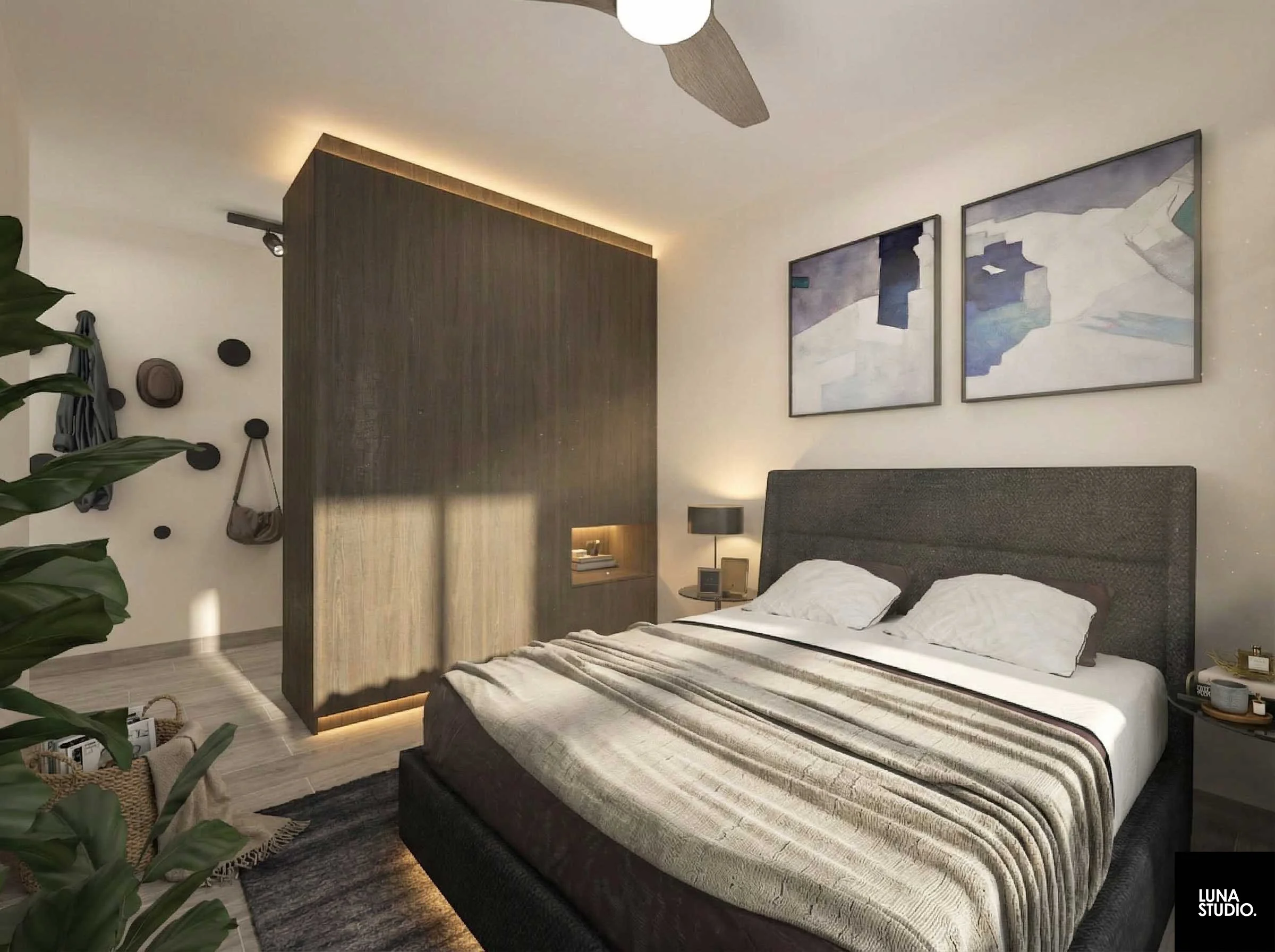 Modern bedroom with a bed that has a gray headboard, white bedding, and two pillows. Beside the bed is a nightstand with a lamp, clock, and small items. Two abstract art pieces hang above the bed. There is a wooden partition on one side with built-in lighting, and hats and bags are hanging on the wall. A plant can be seen in the foreground, and ceiling fan is visible above.