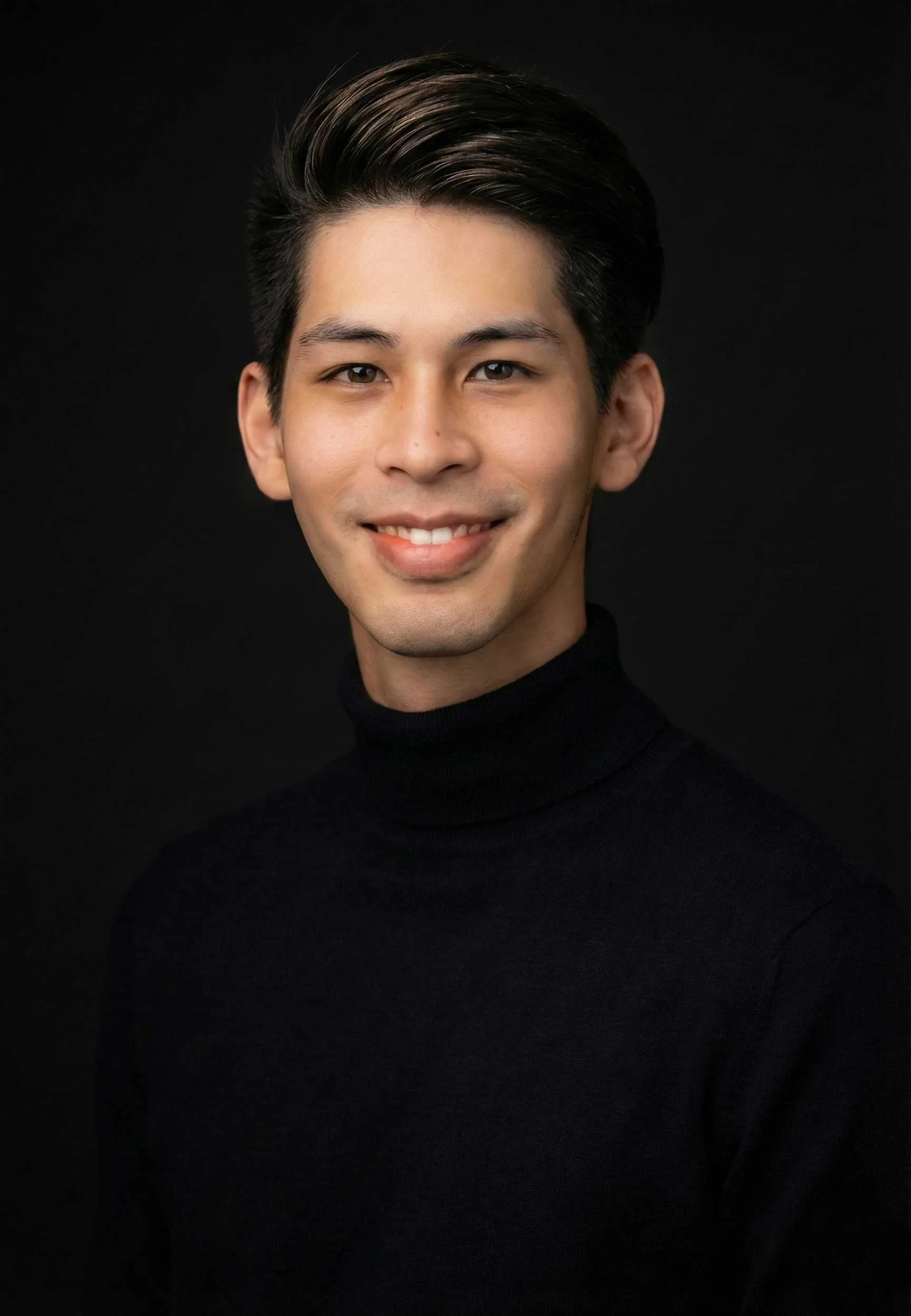 A professional portrait of a young man with dark hair, wearing a black turtleneck, smiling against a black background.
