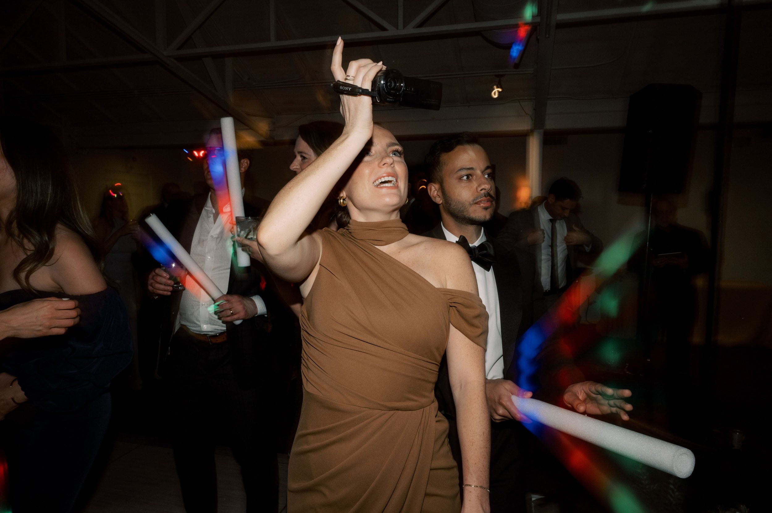 A woman in a brown dress holding a handycam camcorder at a wedding with people waving glow sticks dancing around her.