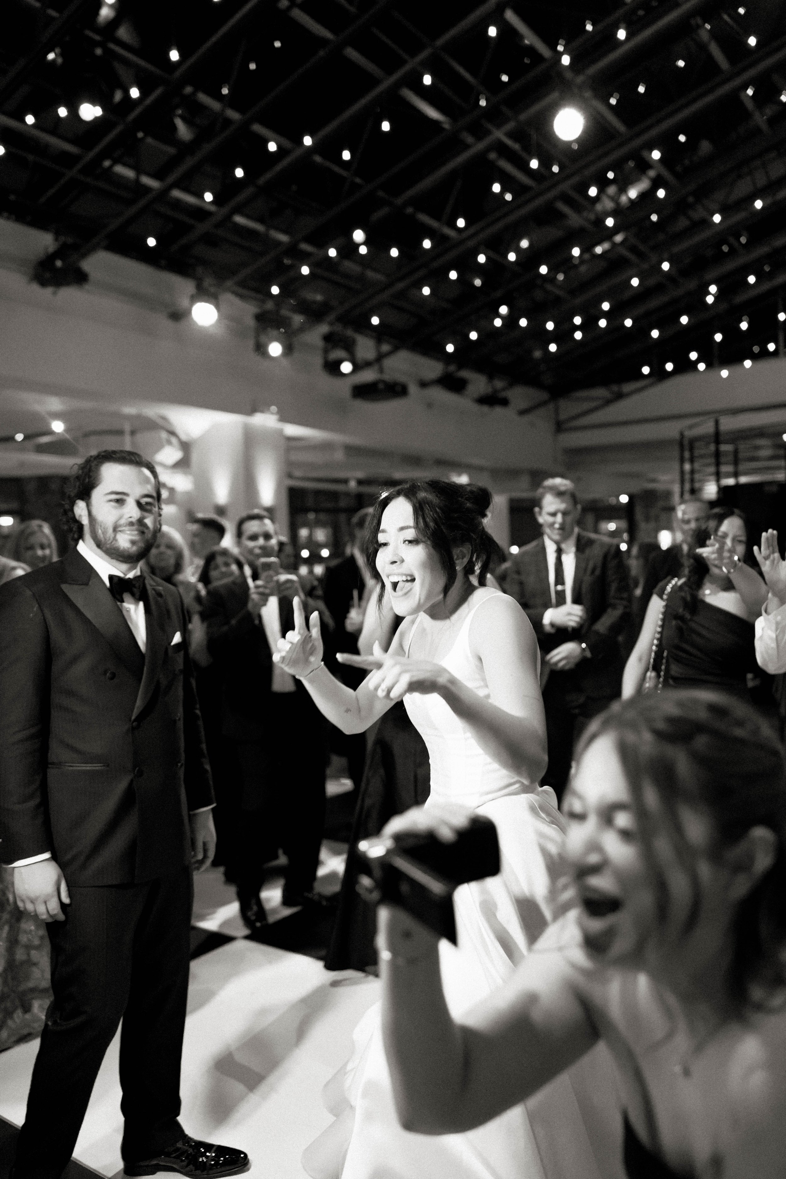 At a wedding, a bride in a white dress is surprised by something, while a man in a tuxedo stands nearby, smiling. A woman in the foreground holds a camcorder. Several people in the background are dressed in formal attire, under a decorated venue.