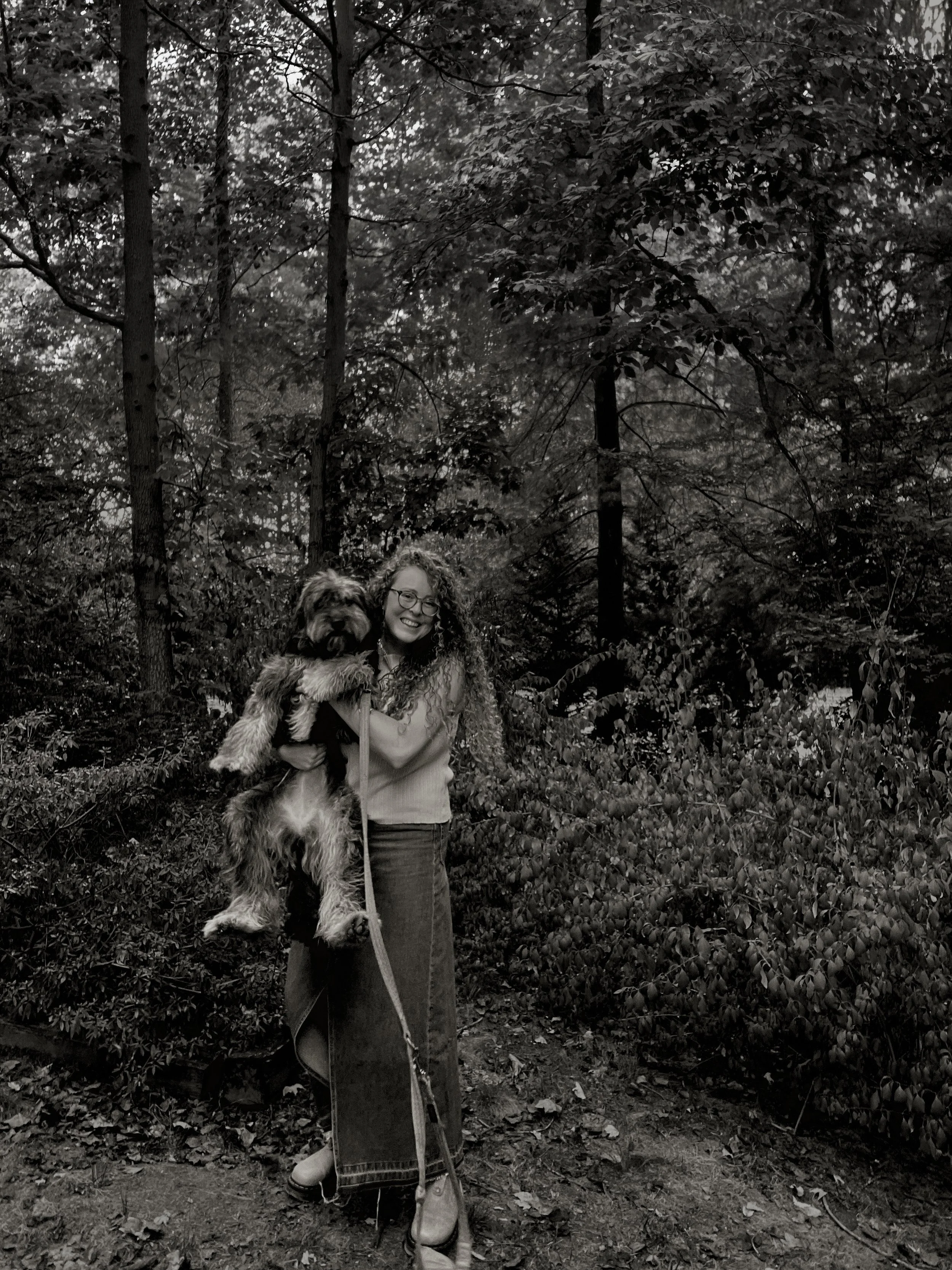 A woman with curly hair and glasses holding a large fluffy dog outdoors in a wooded area.