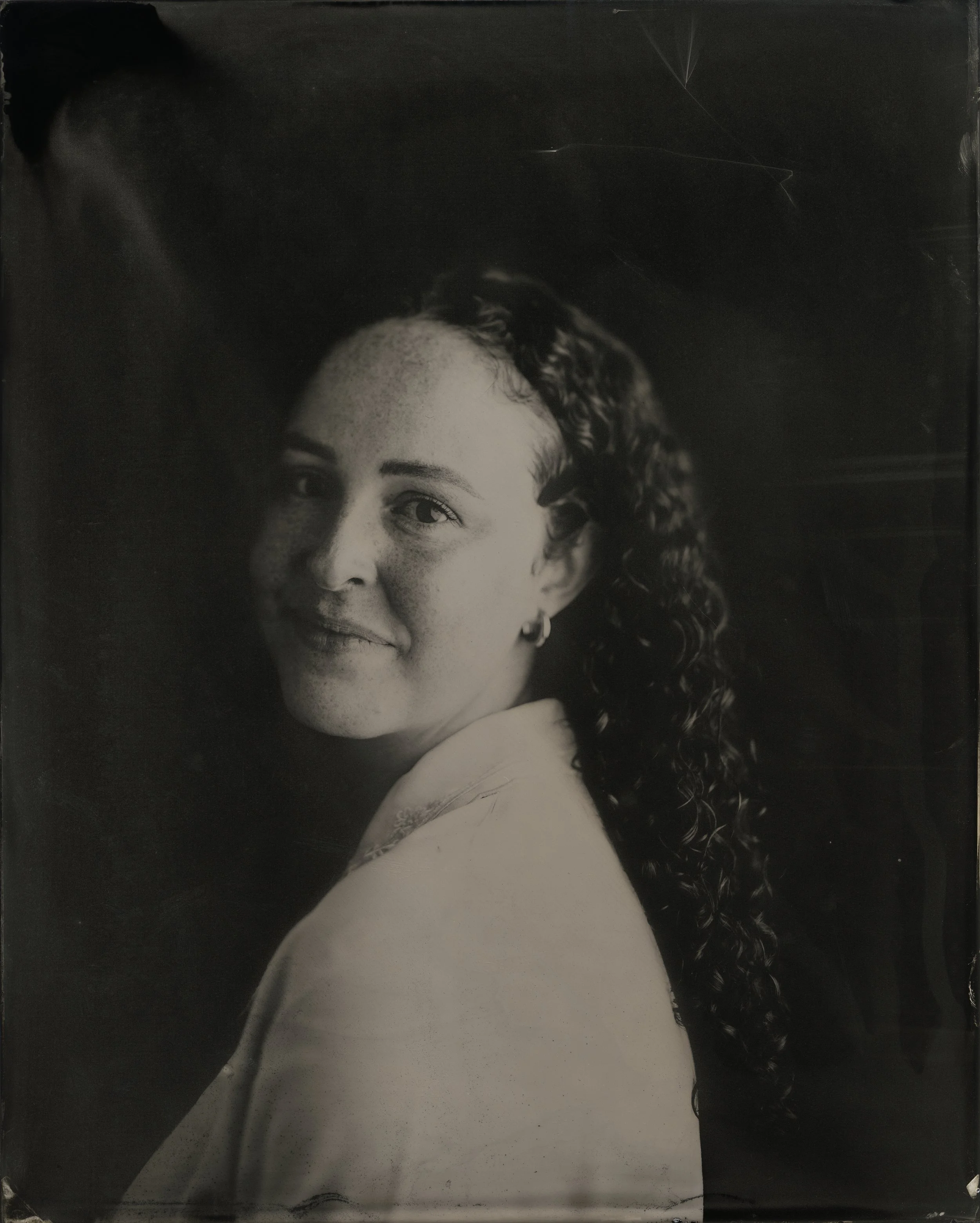 Black and white tintype portrait of a wedding filmmaker with curly hair, smiling softly, wearing a light-colored top and earrings.