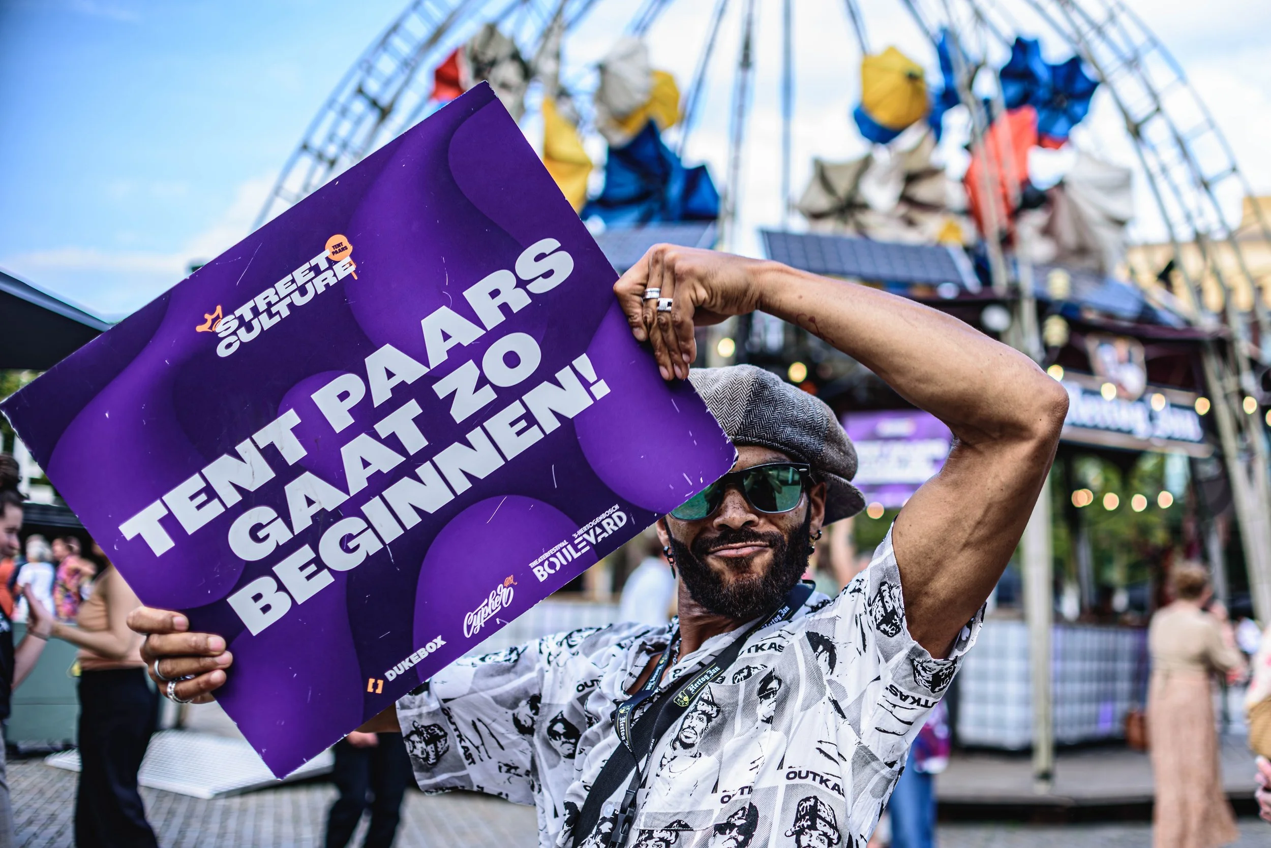 Man met een hoed en zonnebril houdt een paars bord met de tekst 'Tent Paars Gaat Zo Beginnen!' omhoog bij Theaterfestival Boulevard.