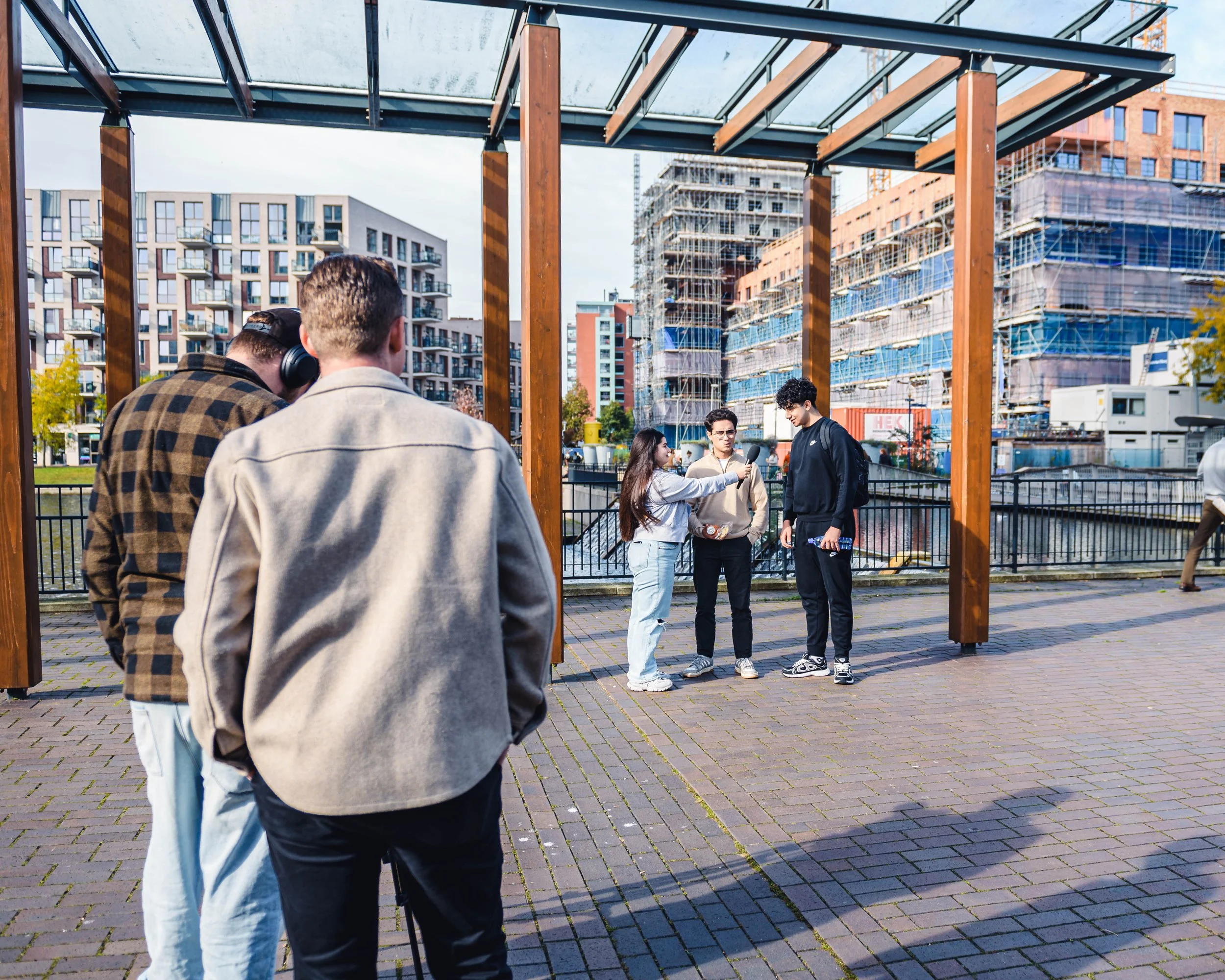 Vijf jonge mensen staan in een stadse omgeving, met een jonge vrouw die een interview afneemt met twee jongens. In de achtergrond zien we gebouwen onder constructie en modern appartementencomplexen.