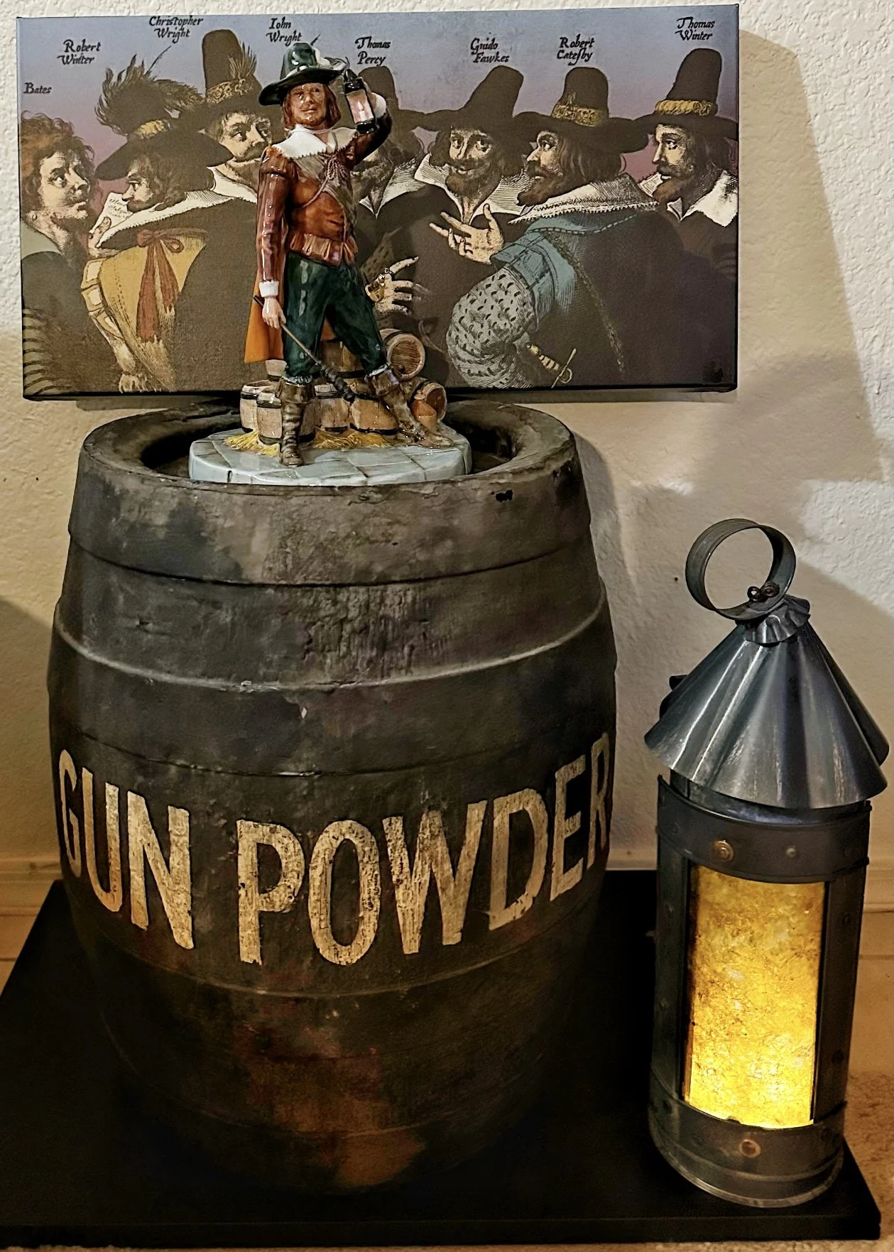 Statue of a man in historic attire on a gunpowder barrel beside a lantern, with a painting of historical figures in the background.