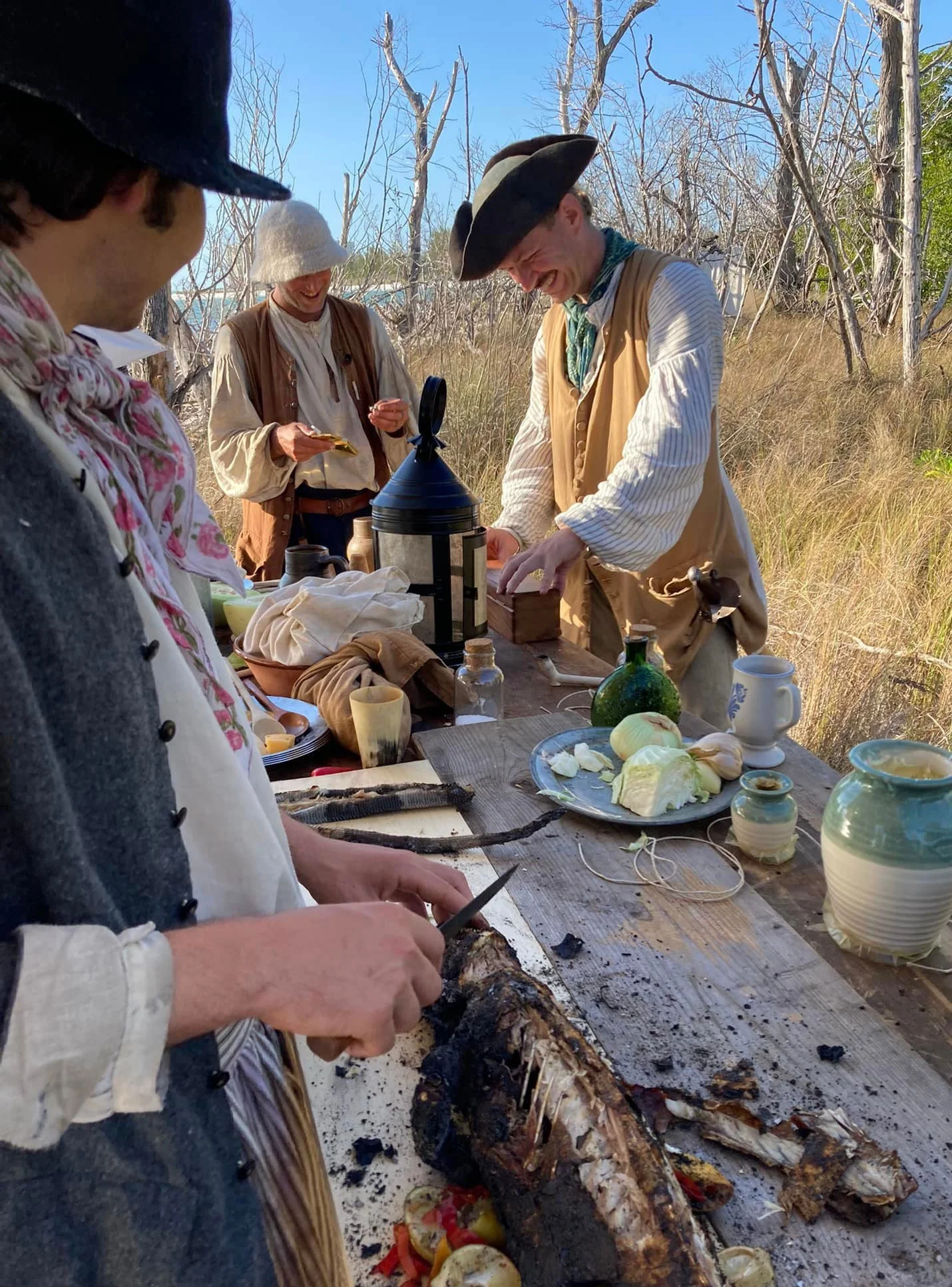 Group of people in historical clothing preparing and eating food outdoors on a wooden table.