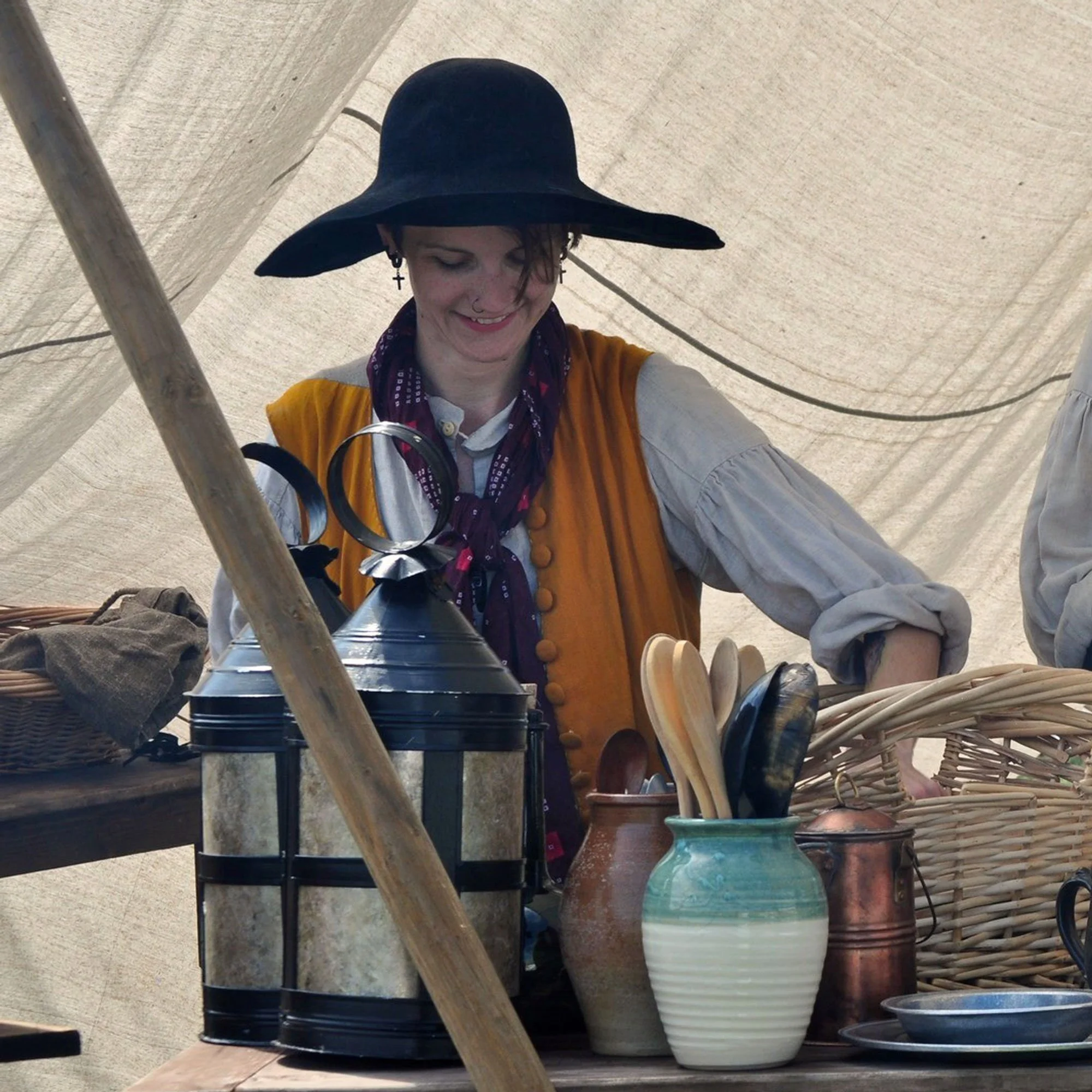 A person wearing a wide-brimmed hat and historical clothing stands behind a table with wooden spoons, ceramic pots, and metal lanterns, set in an outdoor setting.
