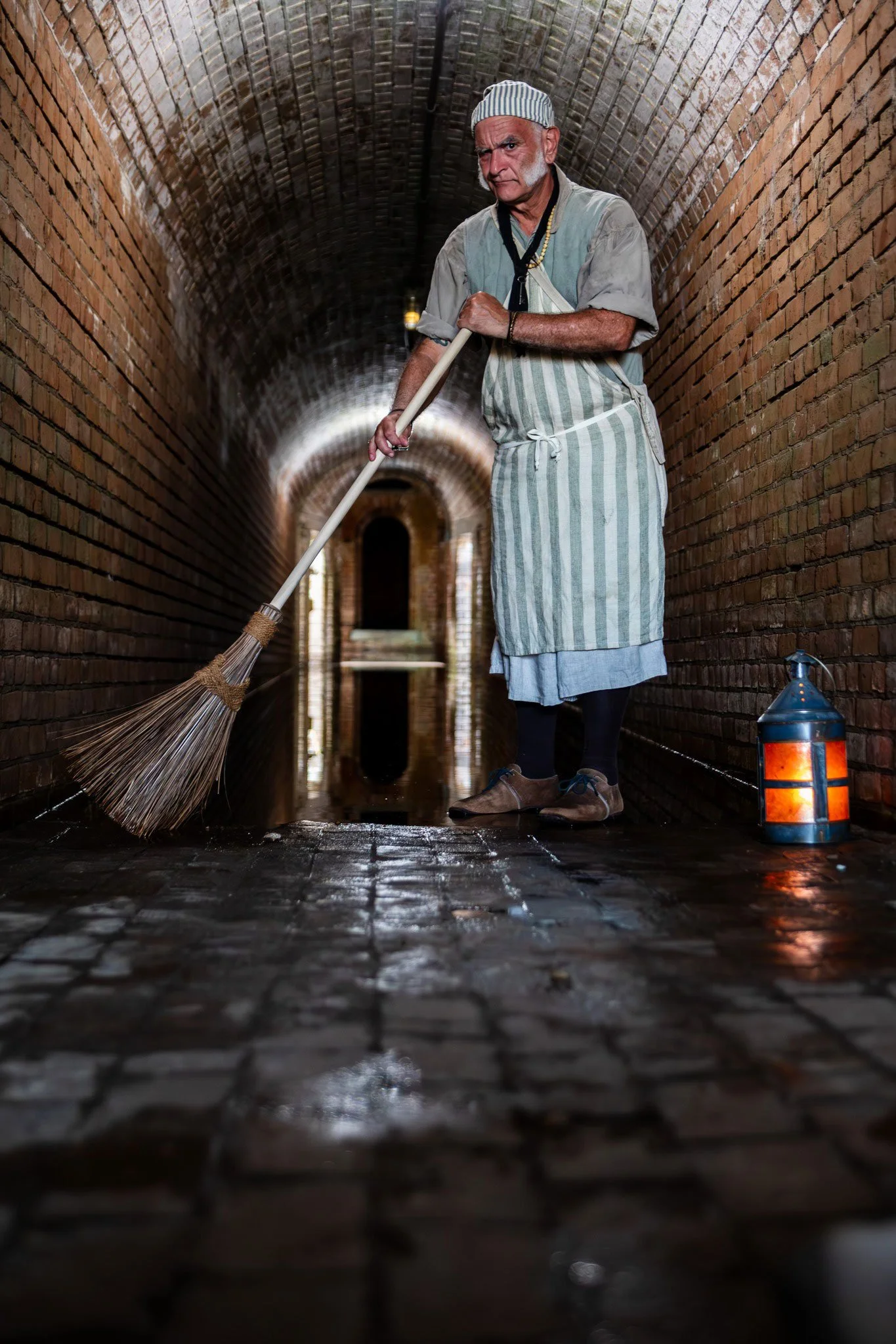 A person dressed in period clothing, including a striped apron and cap, sweeps a wet stone floor in a dimly lit brick tunnel. A lantern with a warm glow is on the ground nearby. The scene suggests a historical or vintage setting.