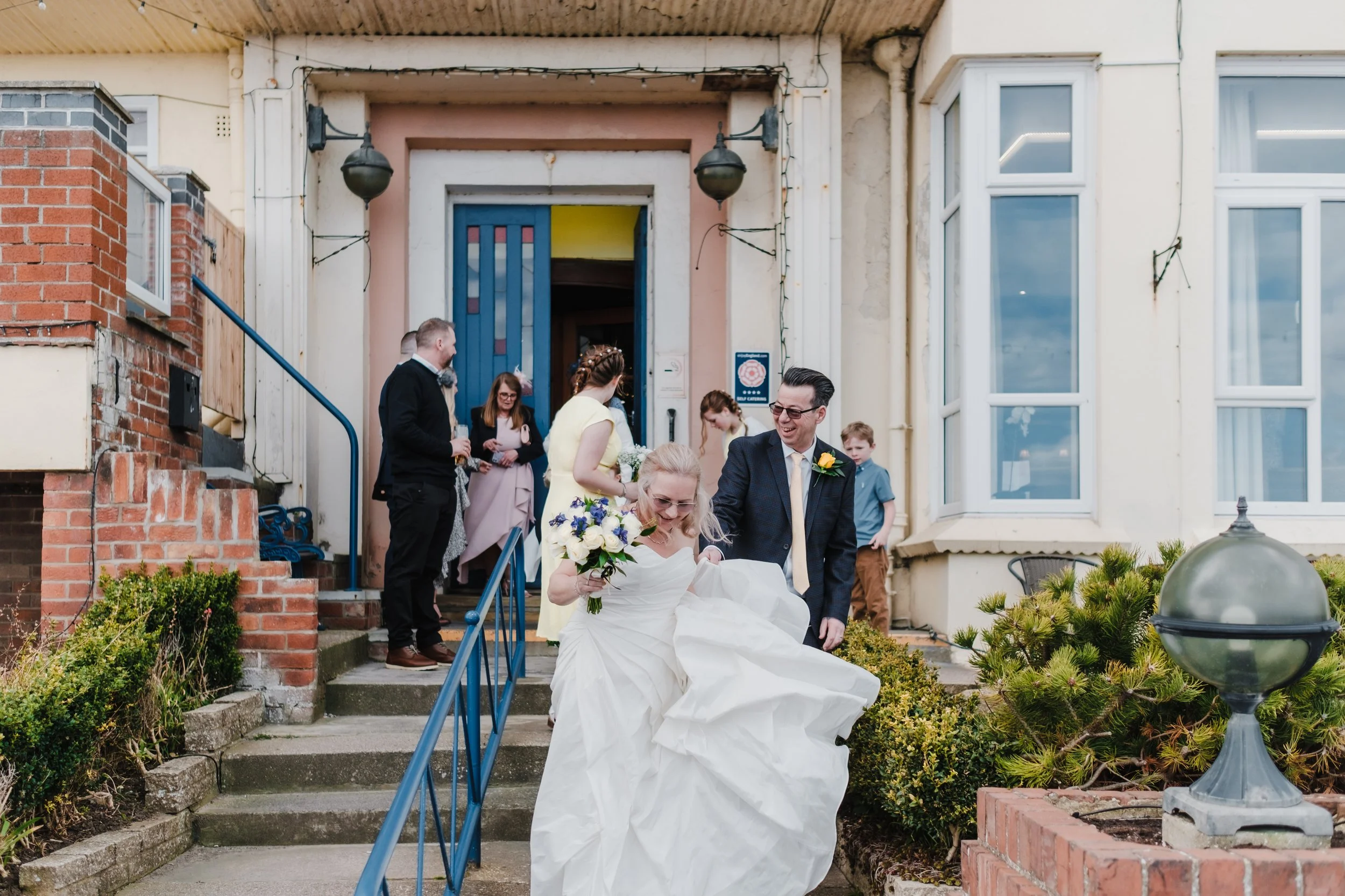 wide shot of bride and groom stepping out of wedding venue, followed by the wedding party