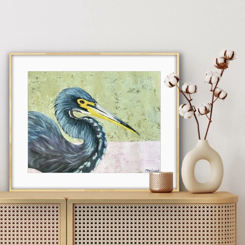 Painting of a heron bird with a yellow and black beak, set against a pastel green and pink background, displayed on a wooden shelf with a white vase of cotton plant branches and a small copper container nearby.
