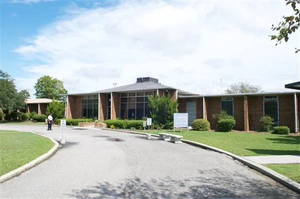 A single-story brick building with large glass windows, surrounded by green grass, bushes, and trees, under a partly cloudy sky.