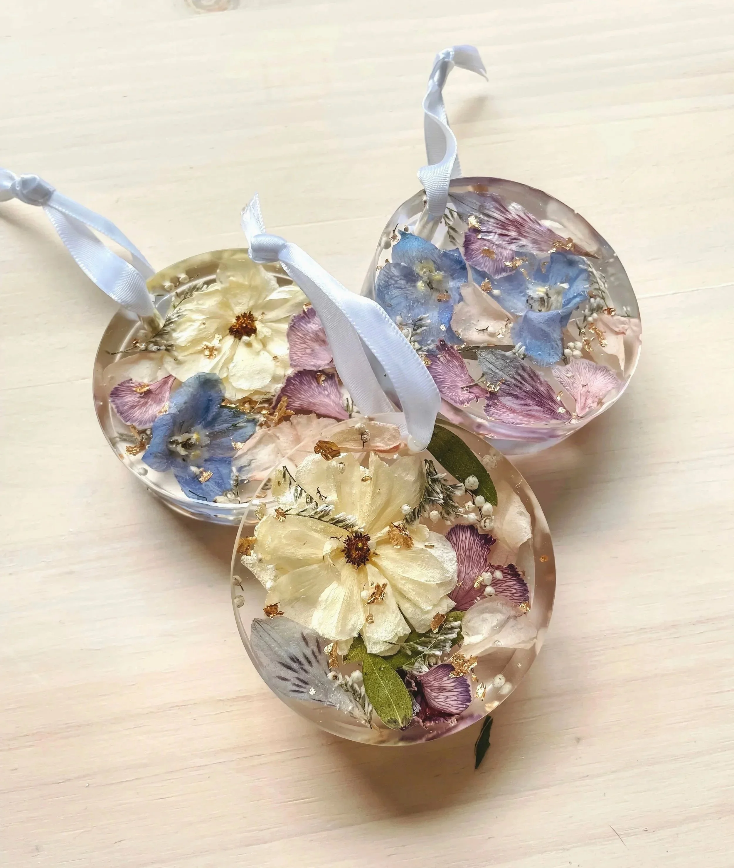 Three clear resin ornaments filled with dried flowers, including blue, pink, and cream petals, with ribbons attached for hanging, placed on a light wooden surface.
