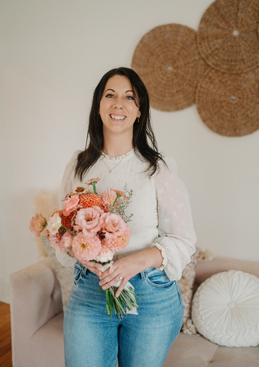 A woman with black hair smiling and holding a pink flower bouquet in a cozy room with woven wall decor and cushions on a sofa.