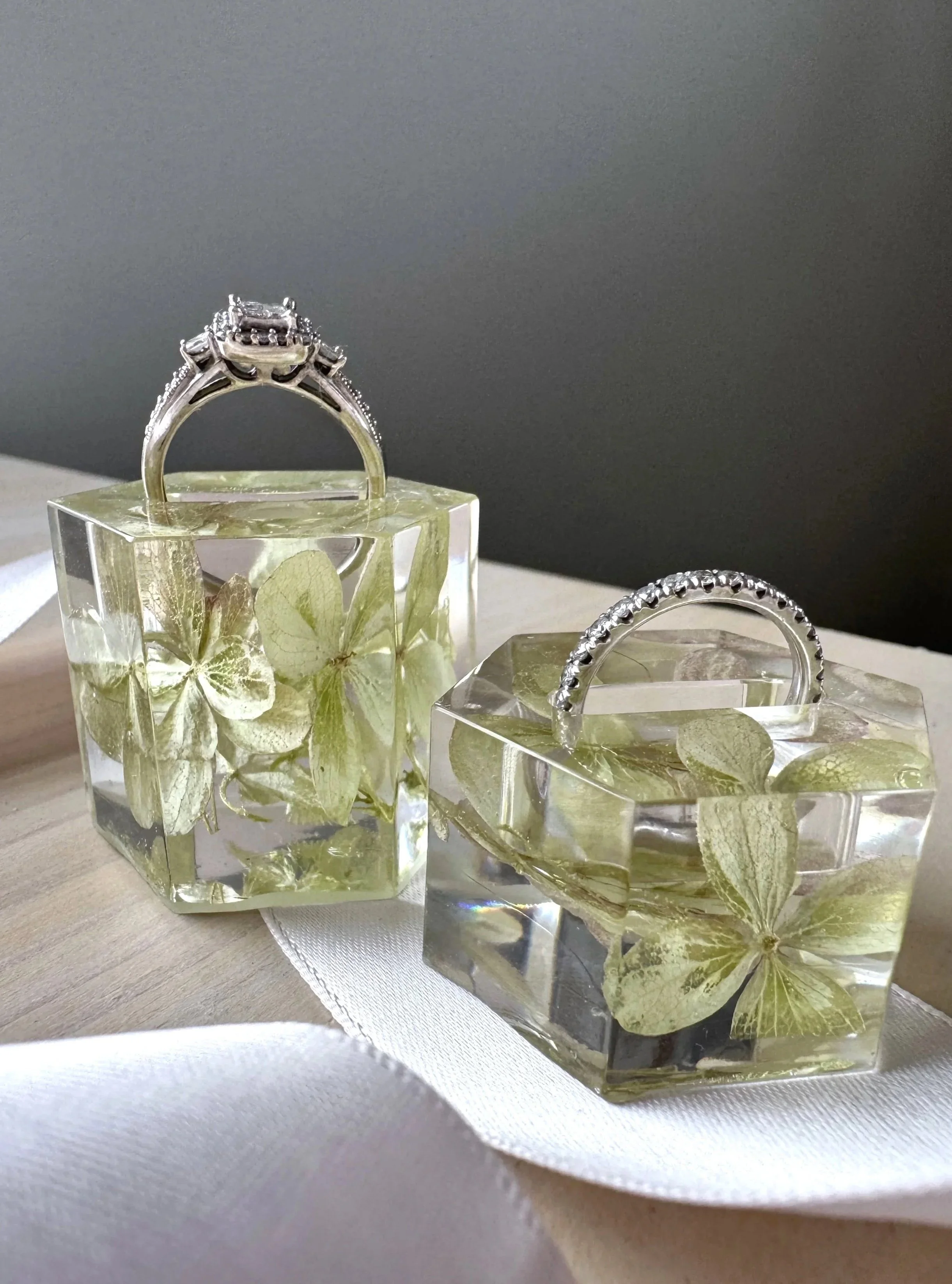 Two transparent resin cubes with preserved pink and greenish hydrangea flowers inside, used as jewelry displays, one holding a gold ring with a pink gemstone and the other a gold ring with pearls, placed on a light-colored surface with a dark shadow background.