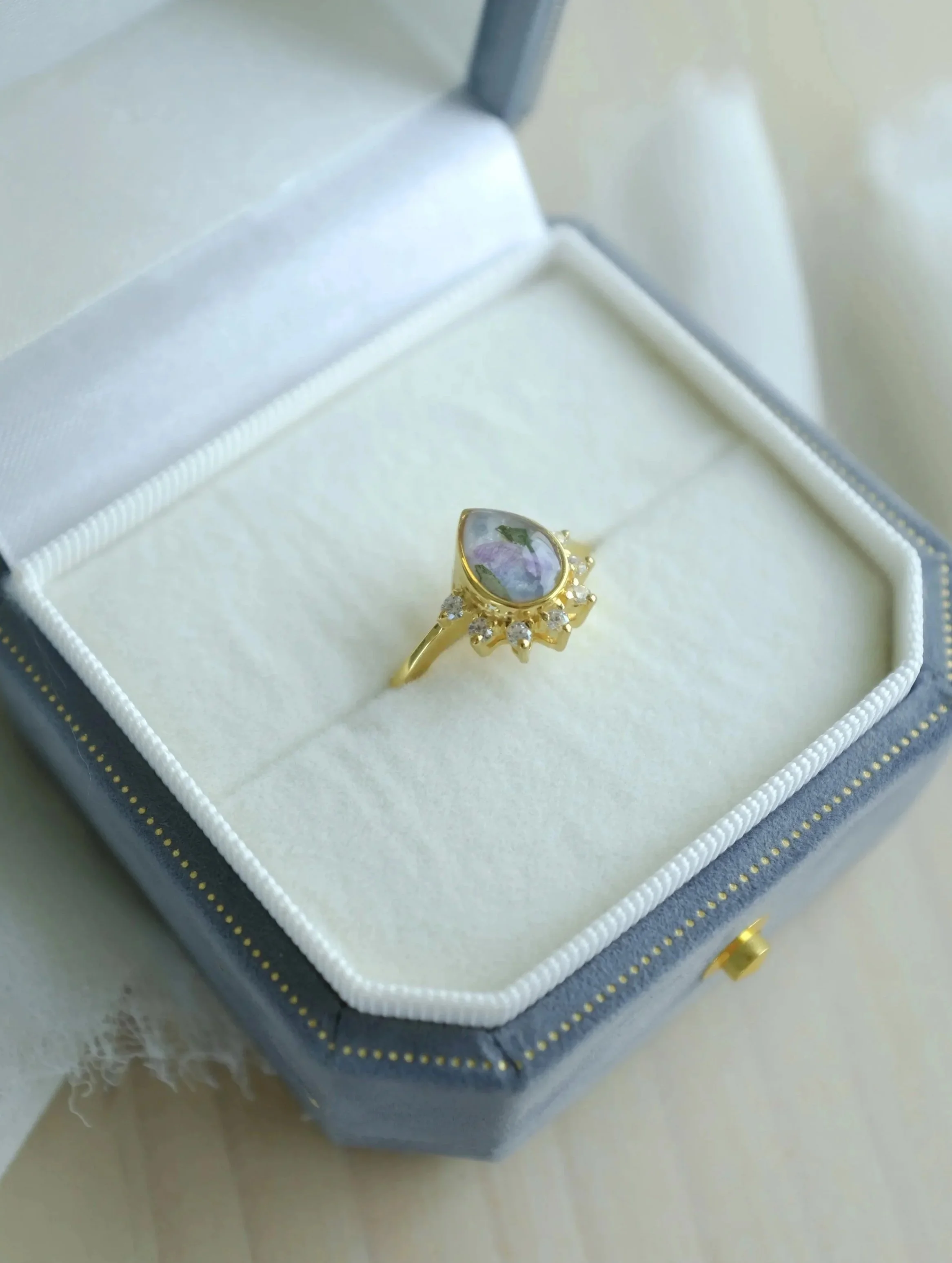 Gold ring with a teardrop-shaped floral design and small gemstones, placed in a white jewelry box.