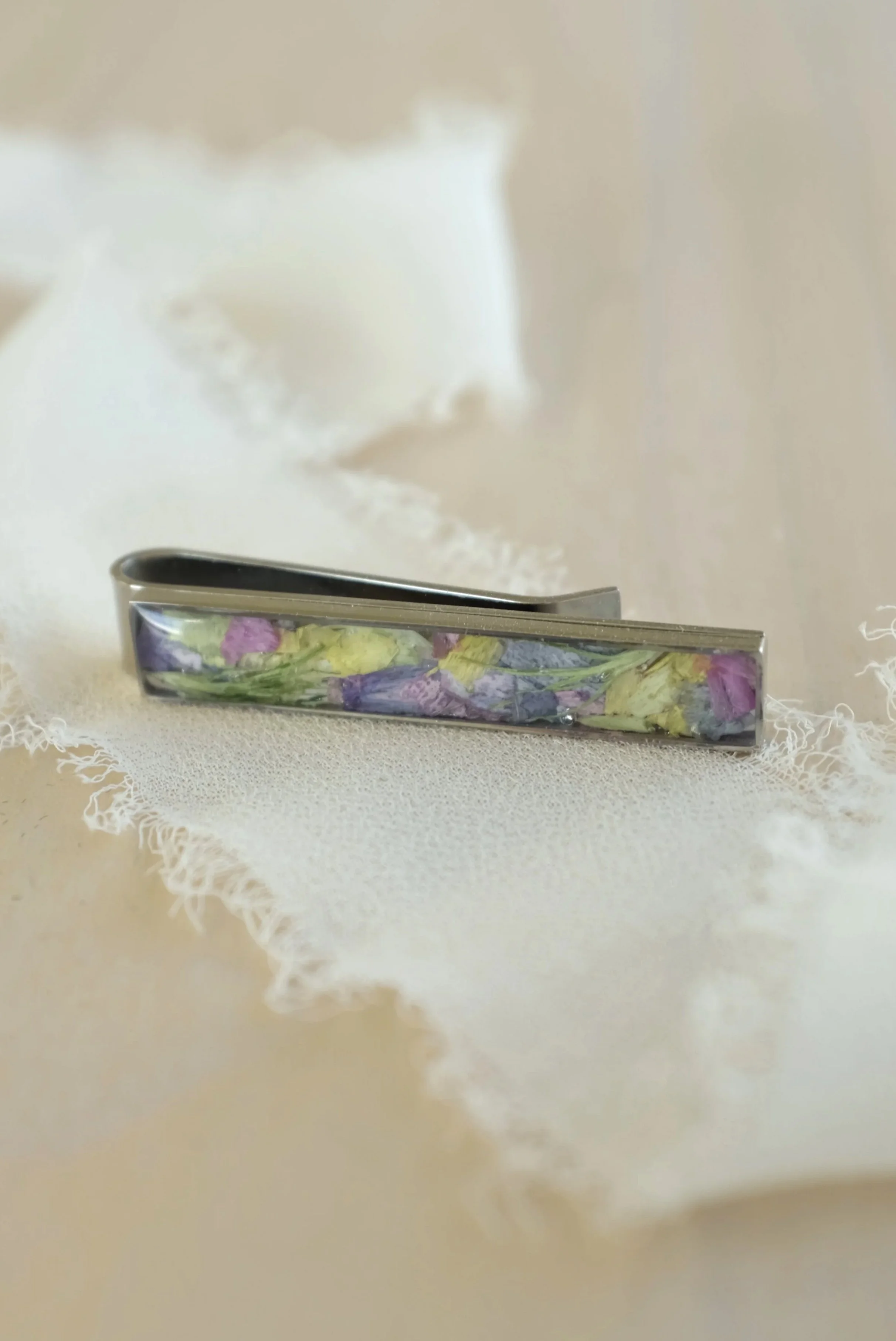 A metal tie clip with embedded dried flower petals in purple, pink, and yellow on a light wooden surface.
