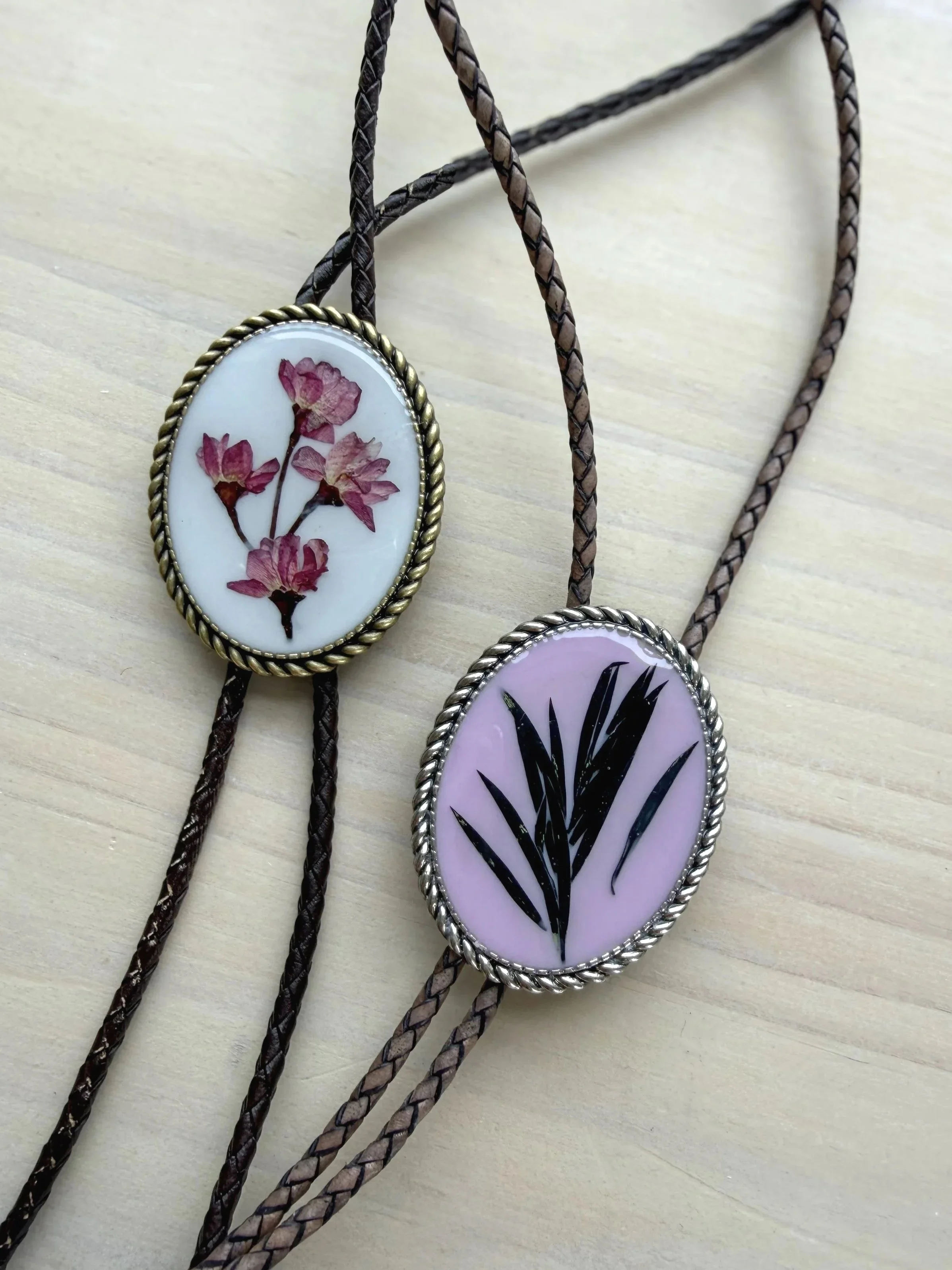 Two braided necklaces with oval pendants. One pendant contains a pressed black and white botanical illustration, and the other contains a pressed golden flower.