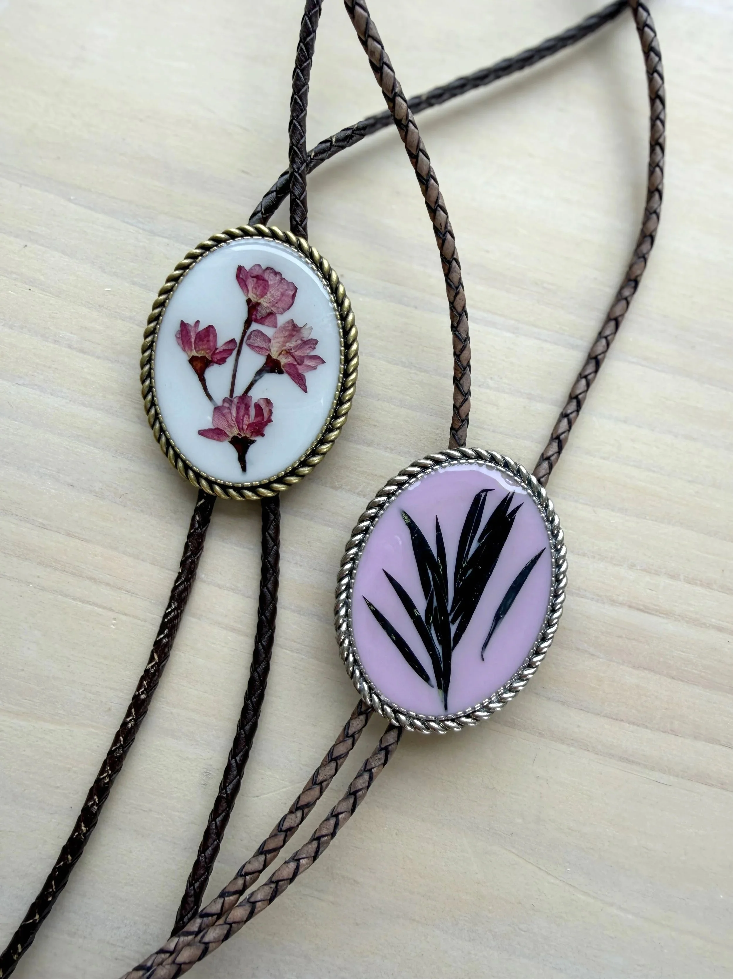 Two braided necklaces with oval pendants. One pendant contains a pressed black and white botanical illustration, and the other contains a pressed golden flower.