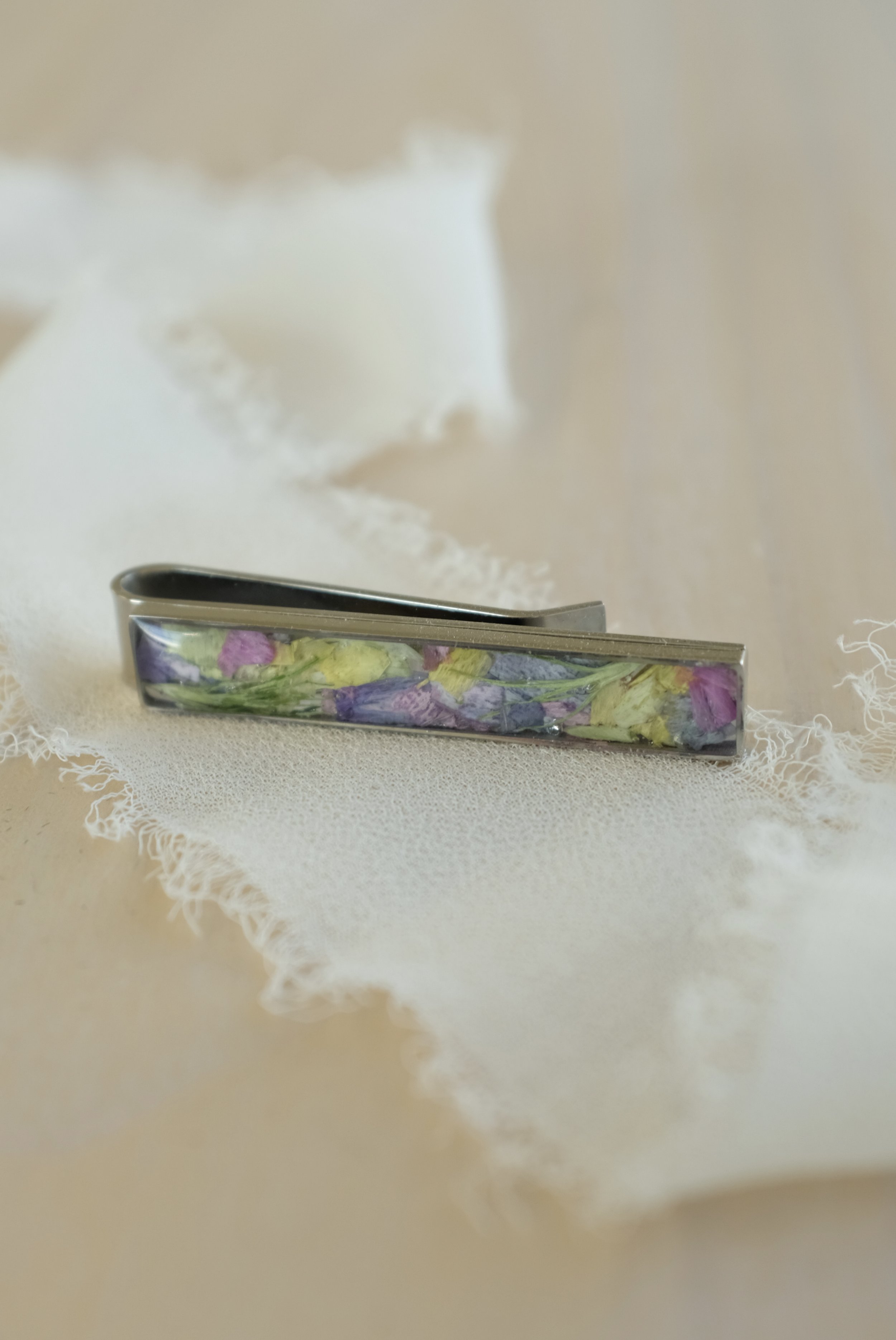 A metal hair clip with embedded dried flower petals in purple, pink, and yellow on a light wooden surface.