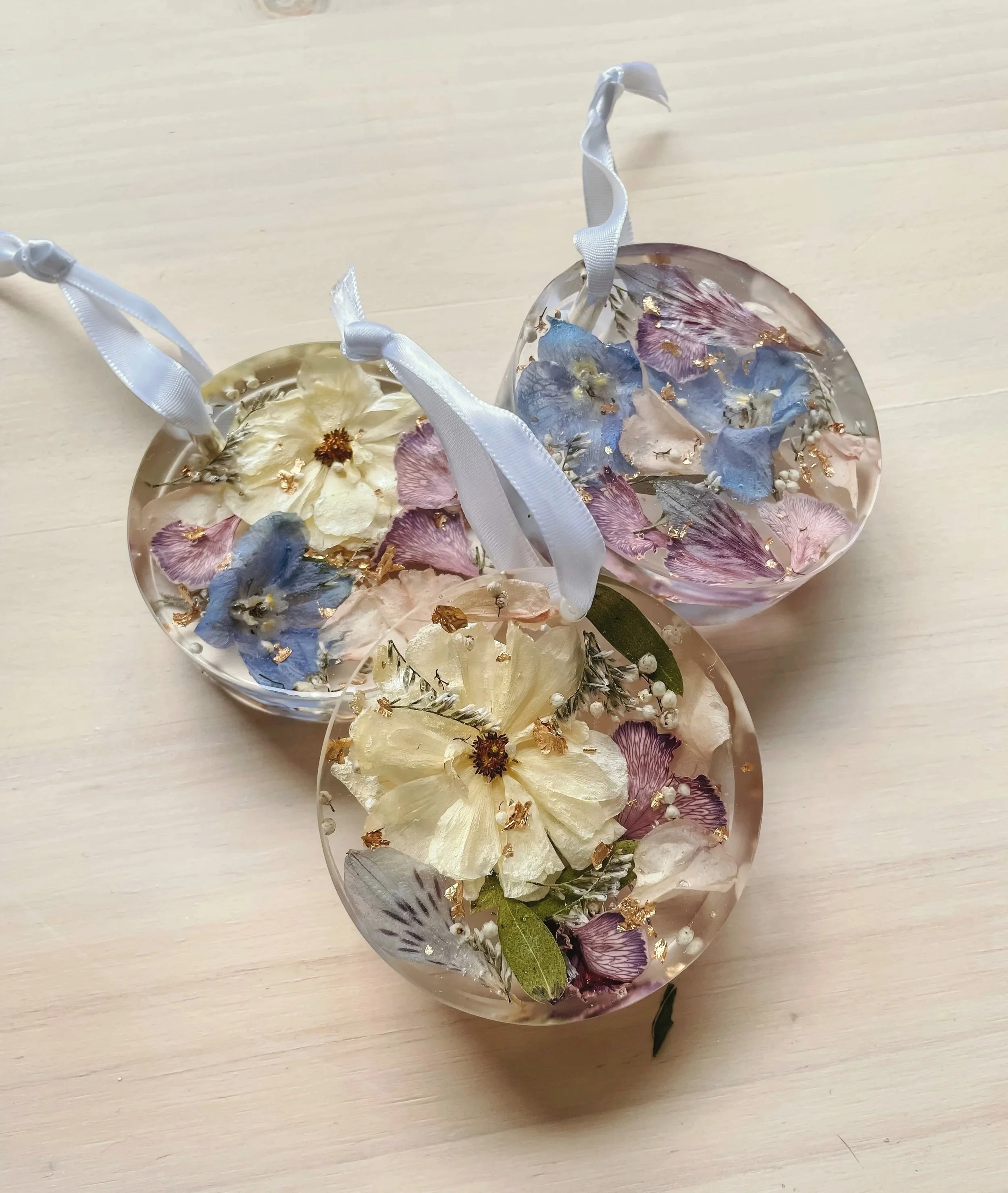 Three clear resin ornaments filled with dried flowers, including blue, pink, and cream petals, with ribbons attached for hanging, placed on a light wooden surface.