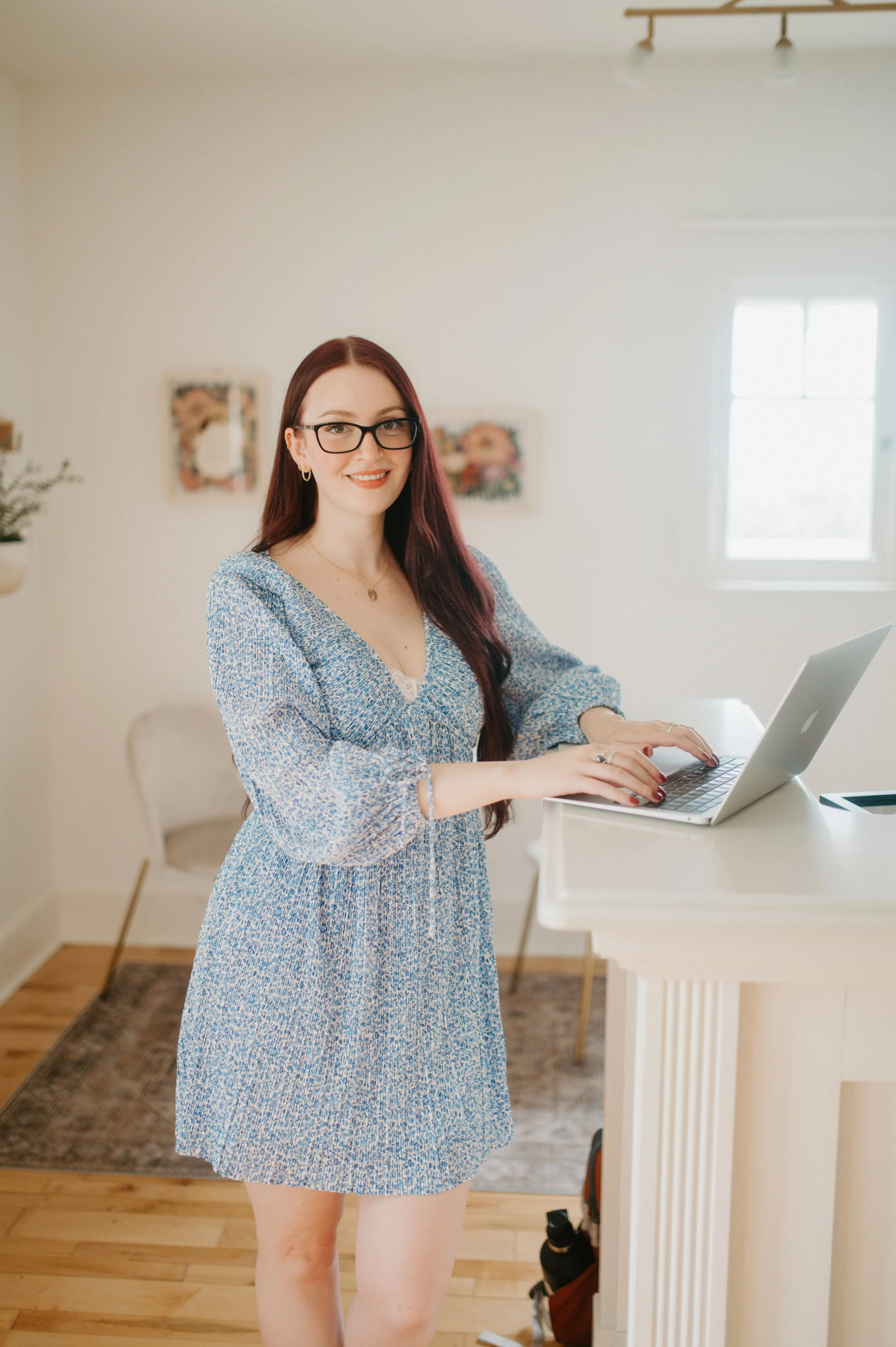 A woman with long dark hair, glasses, and wearing a blue patterned dress working on a laptop in a bright, cozy room.