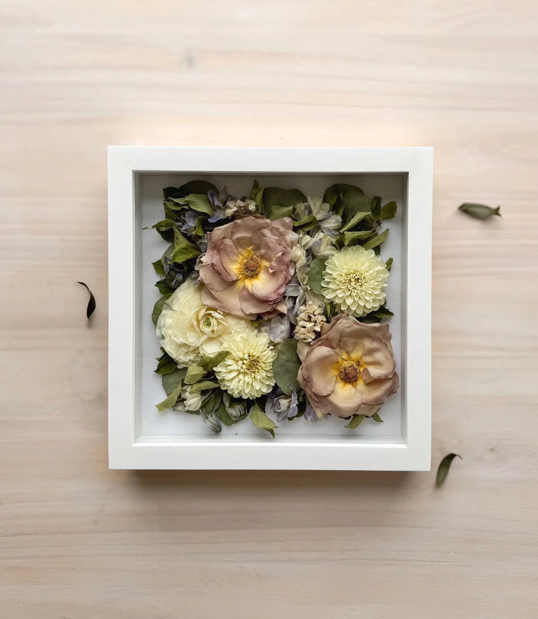 A framed floral arrangement with dried flowers and leaves on a light wooden surface. Some loose leaves are scattered around the frame.