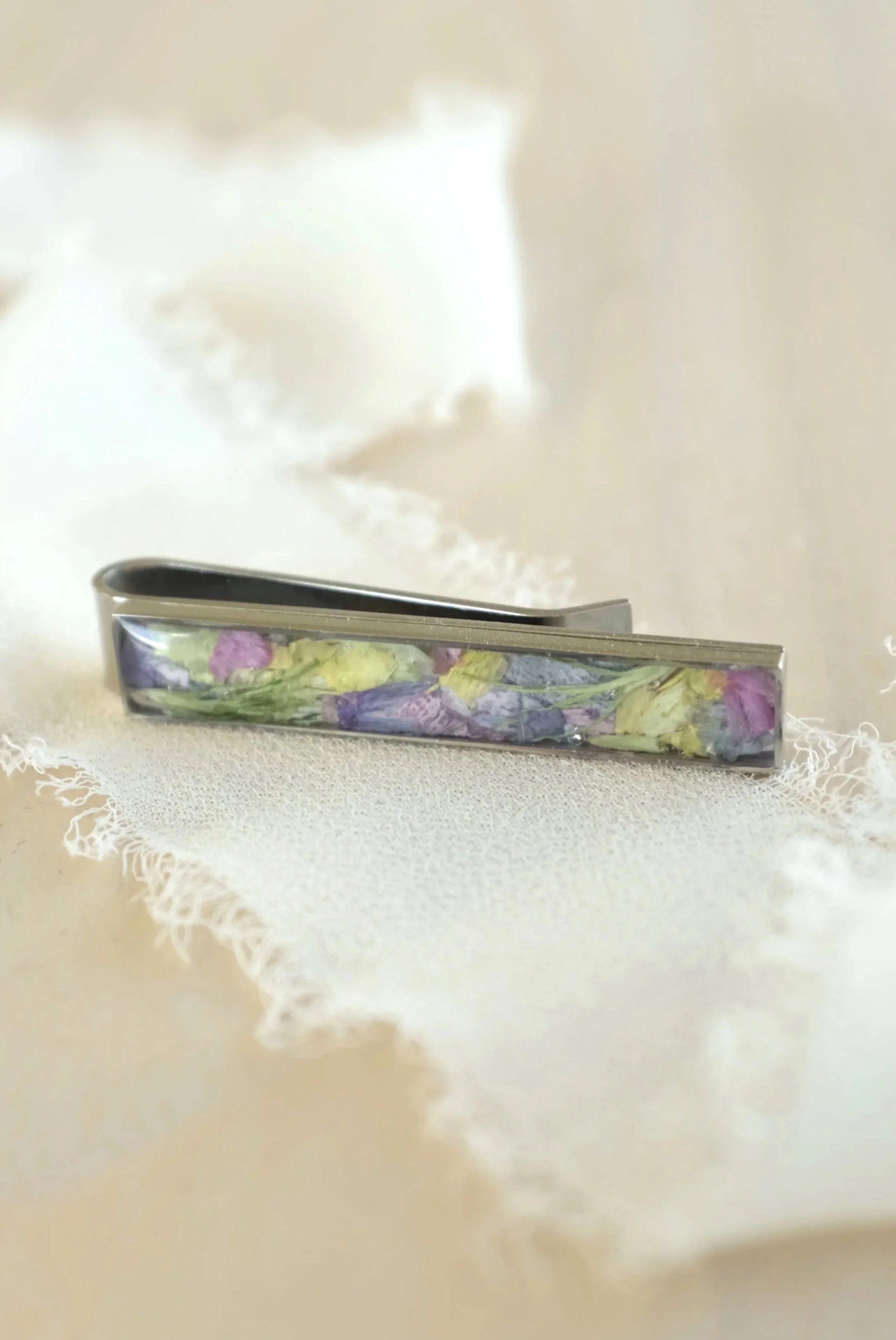 A metal tie clip with embedded dried flower petals in purple, pink, and yellow on a light wooden surface.