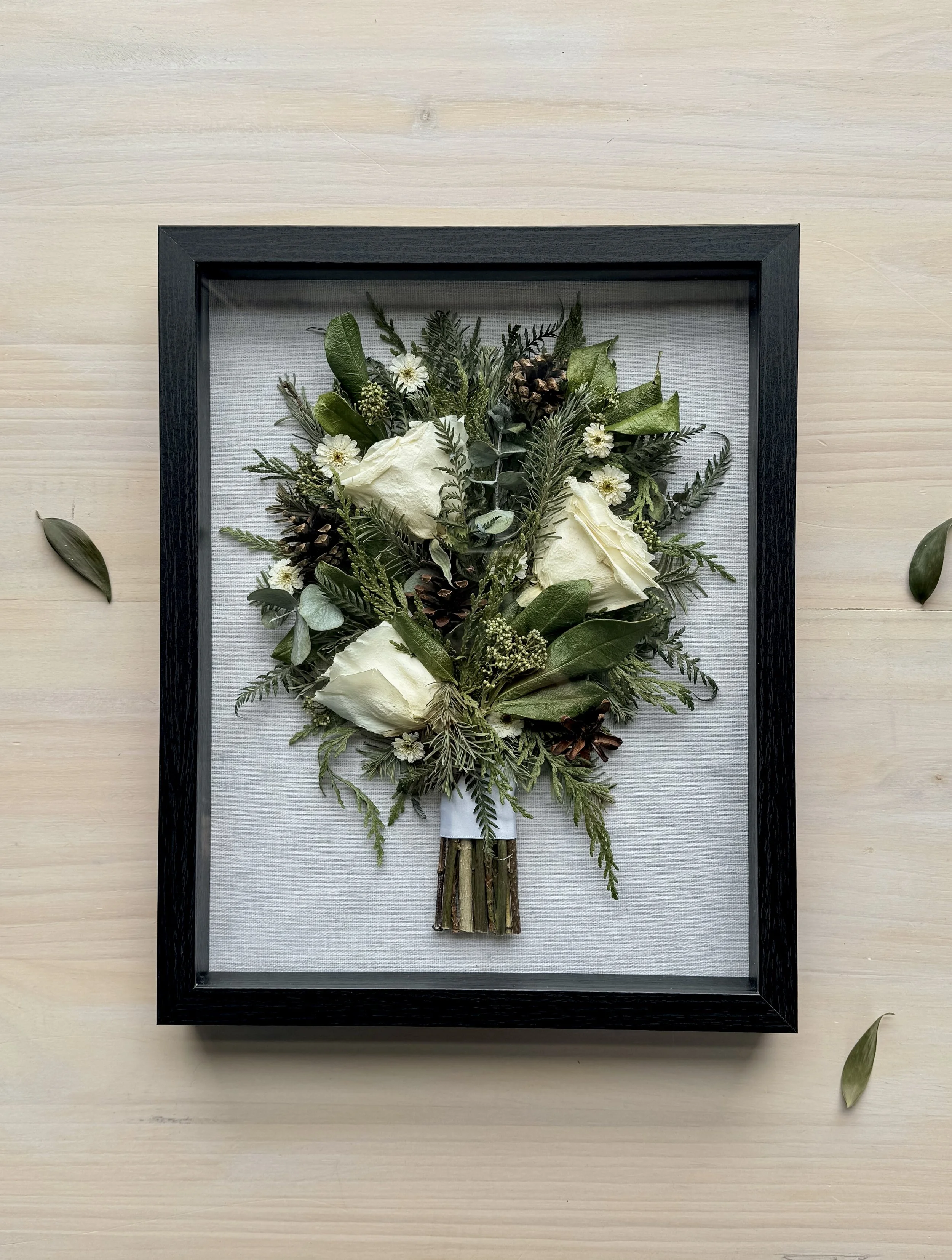 Pressed white roses and greenery arranged as a bouquet in a shadow box frame.