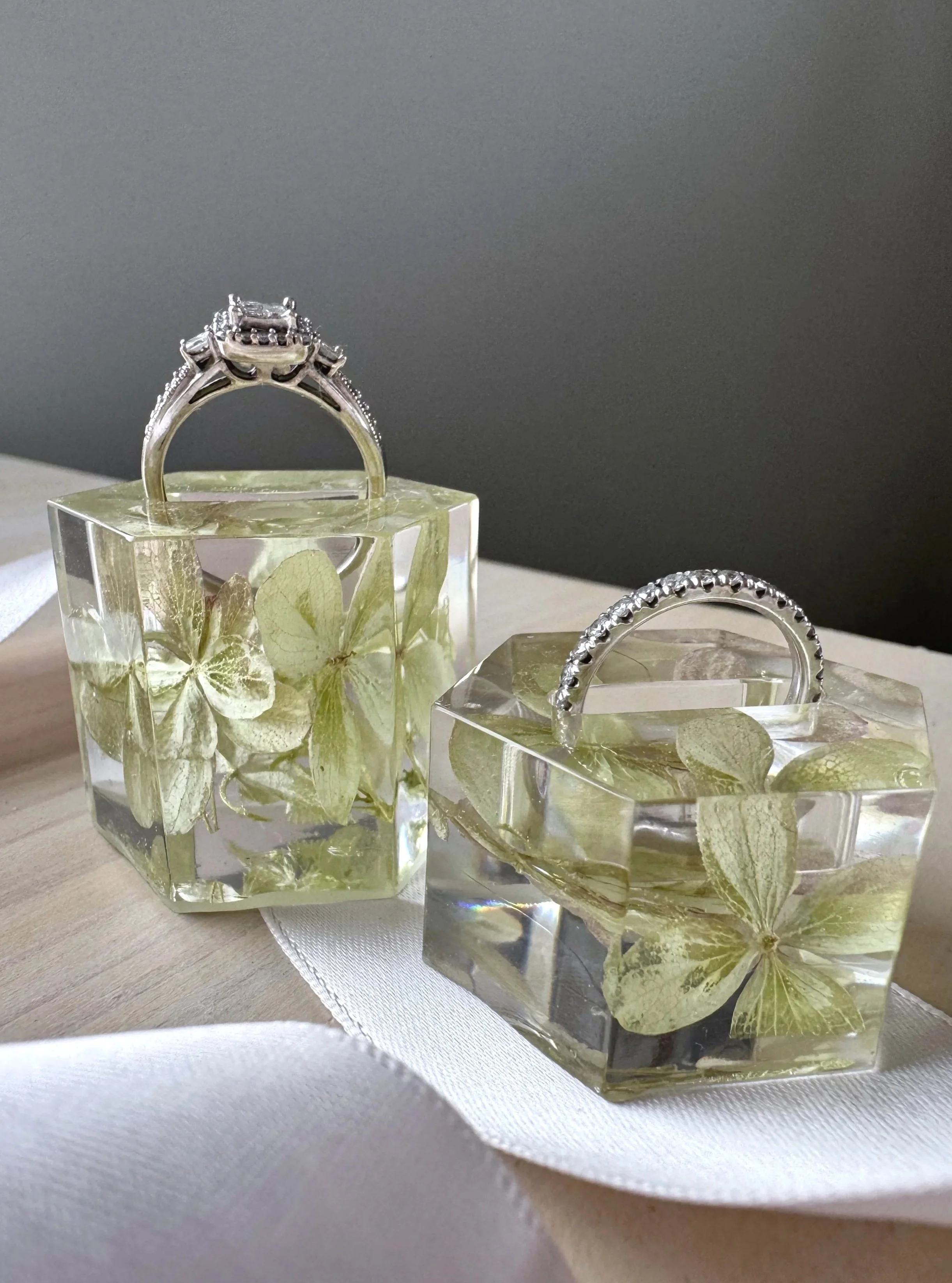 Two transparent resin cubes with preserved pink and greenish hydrangea flowers inside, used as jewelry displays, one holding a gold ring with a pink gemstone and the other a gold ring with pearls, placed on a light-colored surface with a dark shadow background.