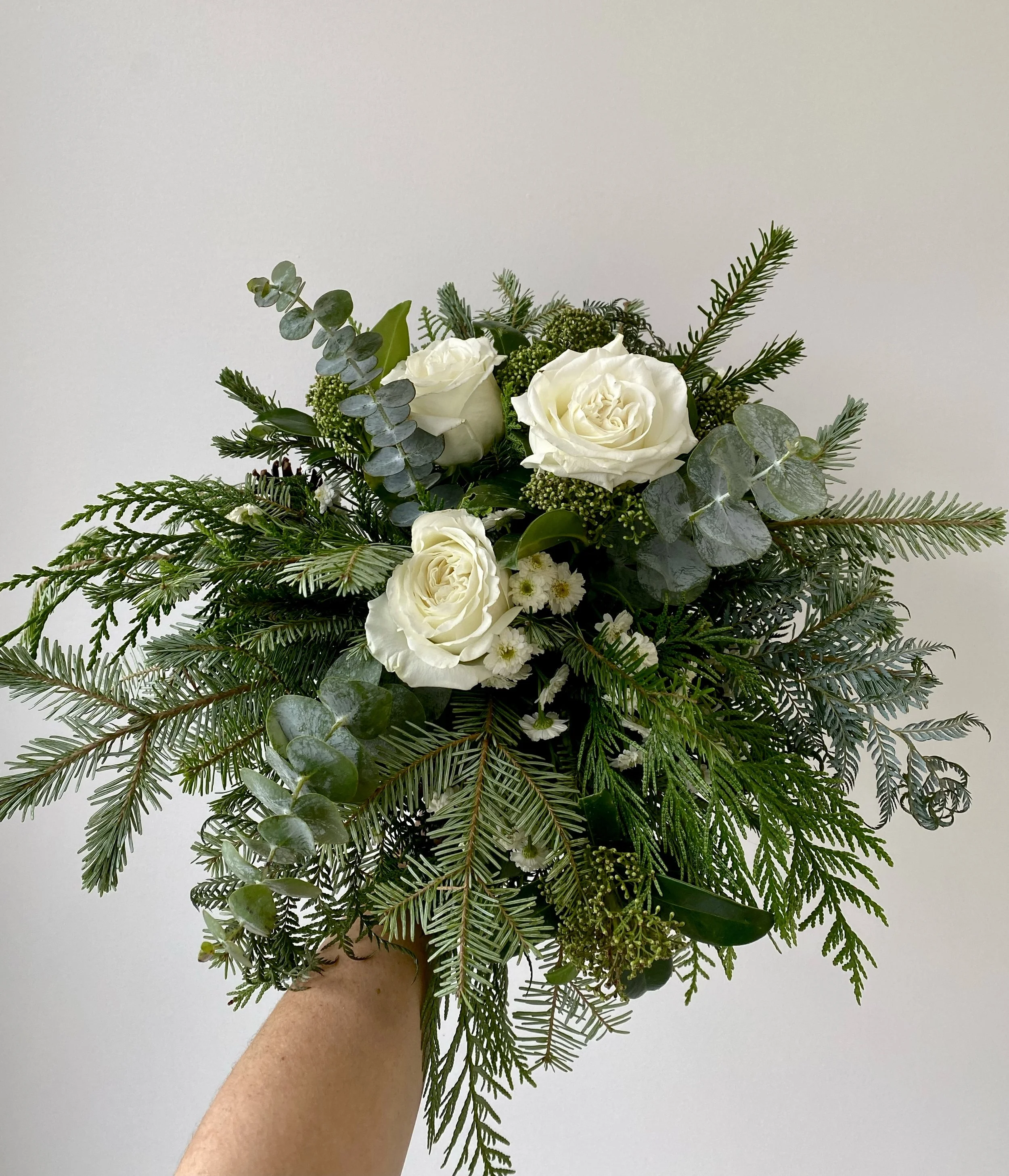 A hand holding a bouquet of white roses, eucalyptus, and various greenery against a plain white wall.