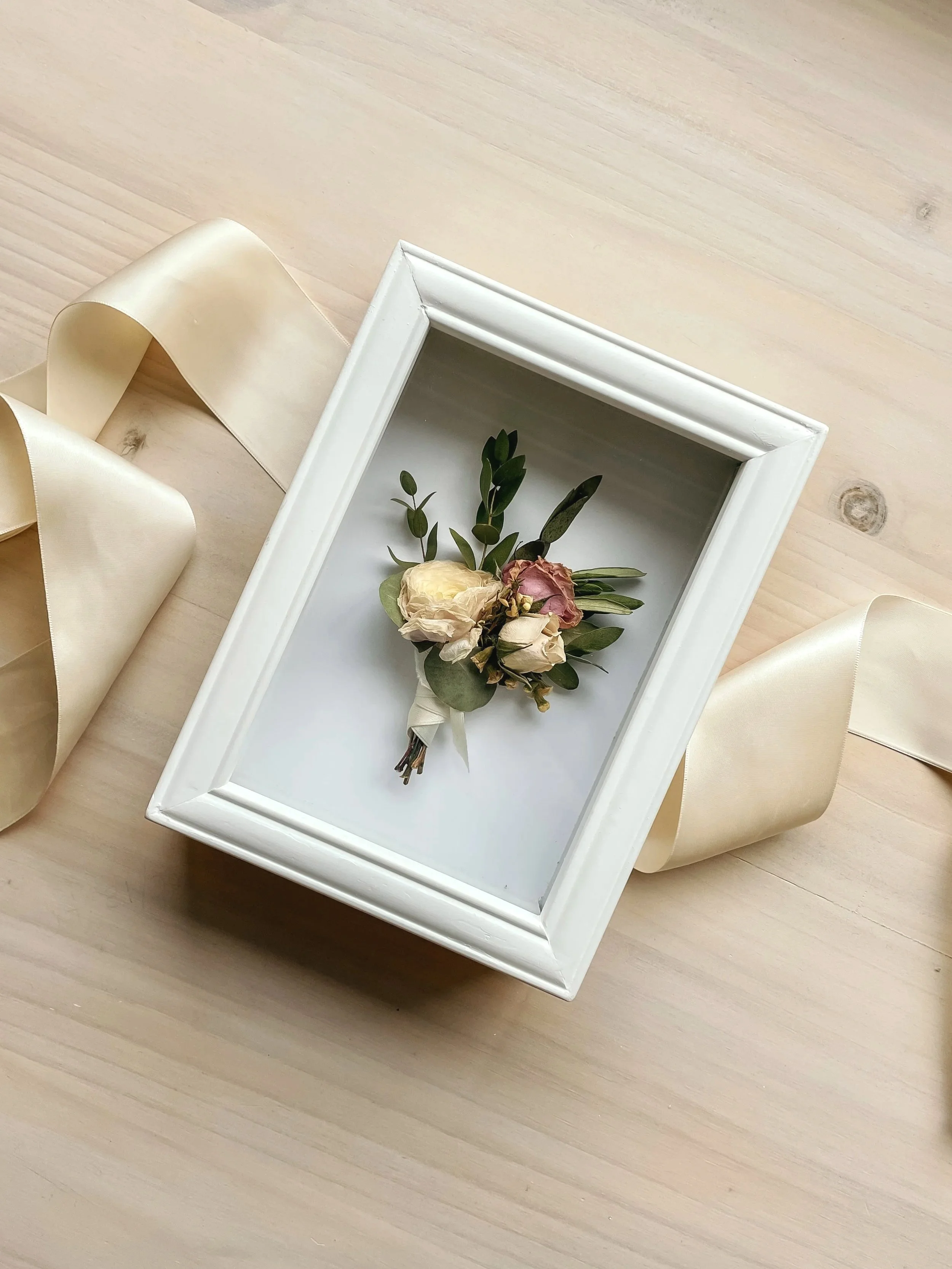 A small bouquet of dried roses and greenery in a white frame on a wooden surface, with a cream-colored satin ribbon nearby.