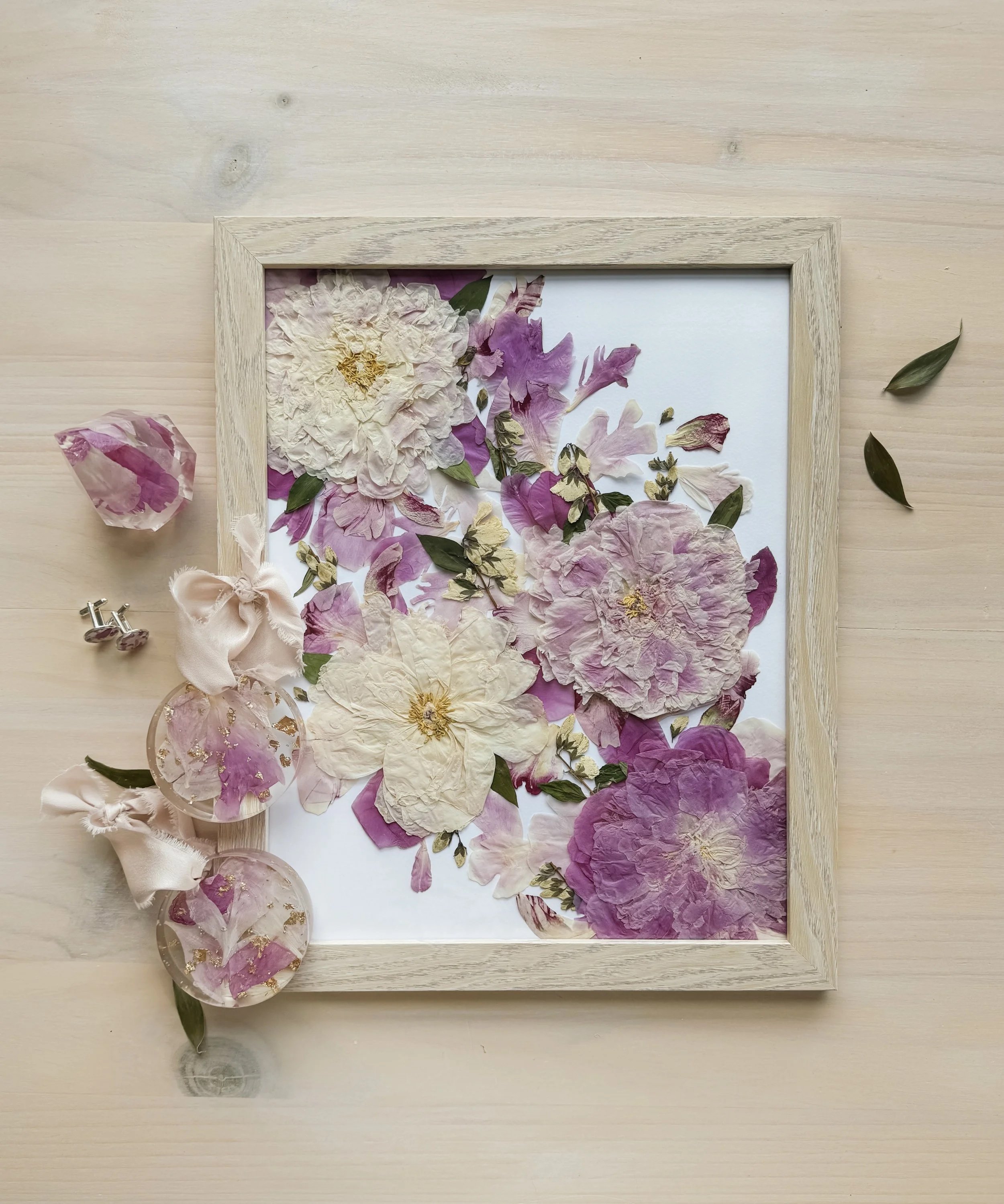 A framed display of pressed pink and white flowers and green leaves on a white background, with a few loose flowers and leaves around it on a light wooden surface.