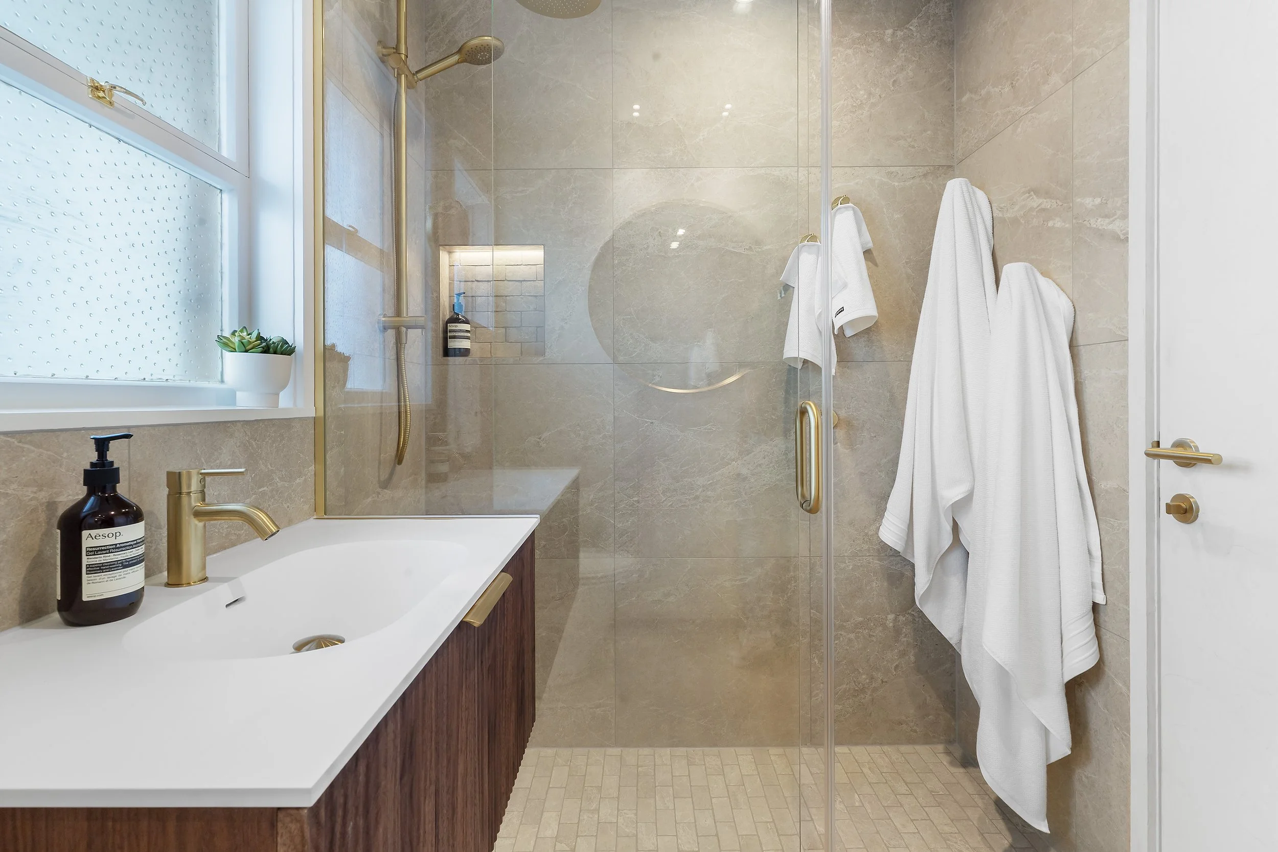 The simple wooden vanity and tower cabinet evoke a driftwood feel, seamlessly blending with the beige, sand-colored tiles and mosaics make you feel like you are stepping into a coastal retreat. This curated mix immediately delivers a calm, beach-insp