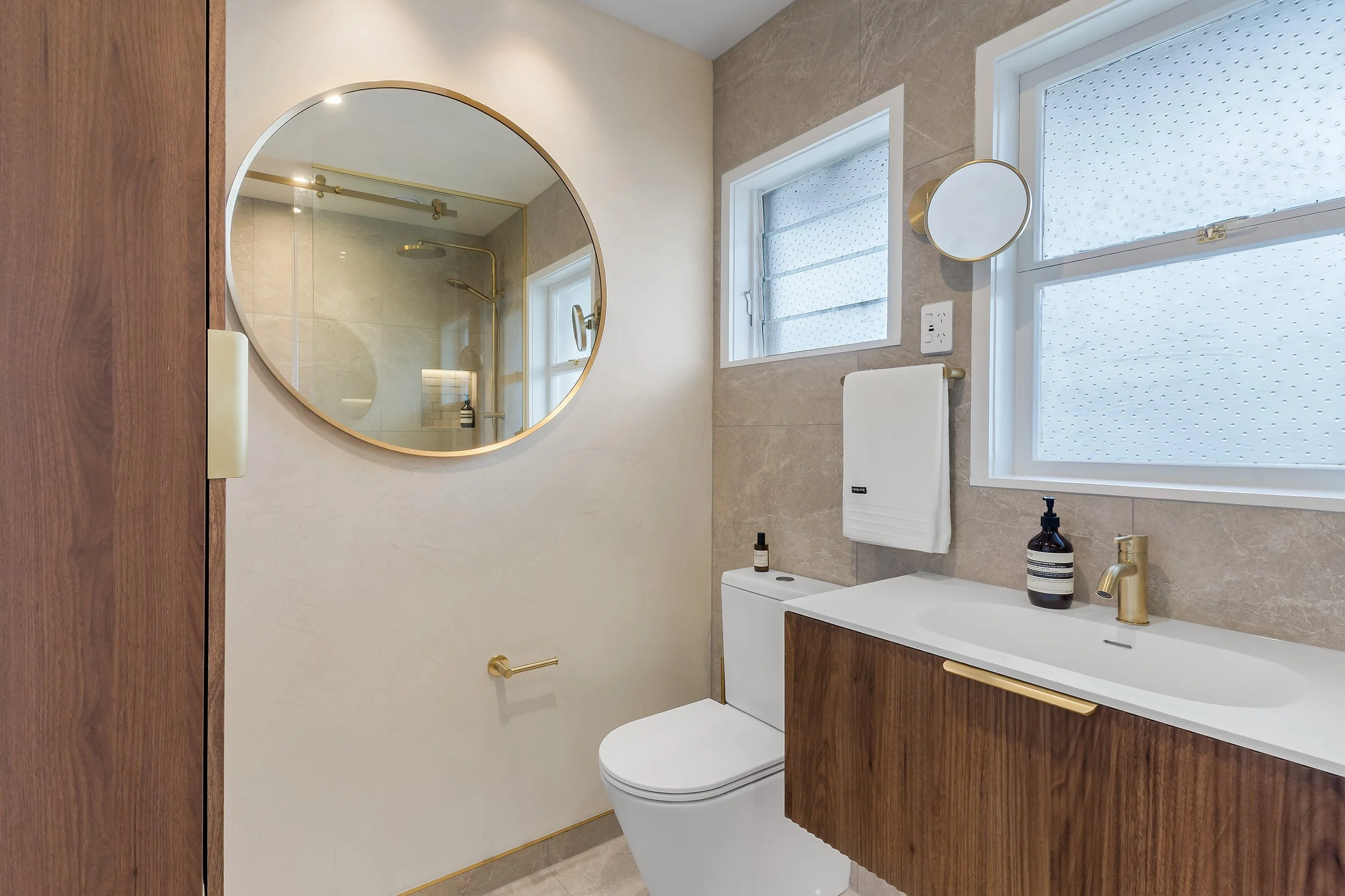 The design is a study in texture and contrast: Matt white finishes and the driftwood-inspired vanity and cabinet flow seamlessly against the beige tiles and fresco- wax finished walls and ceiling. Throughout the space, brushed brass finishes introduc