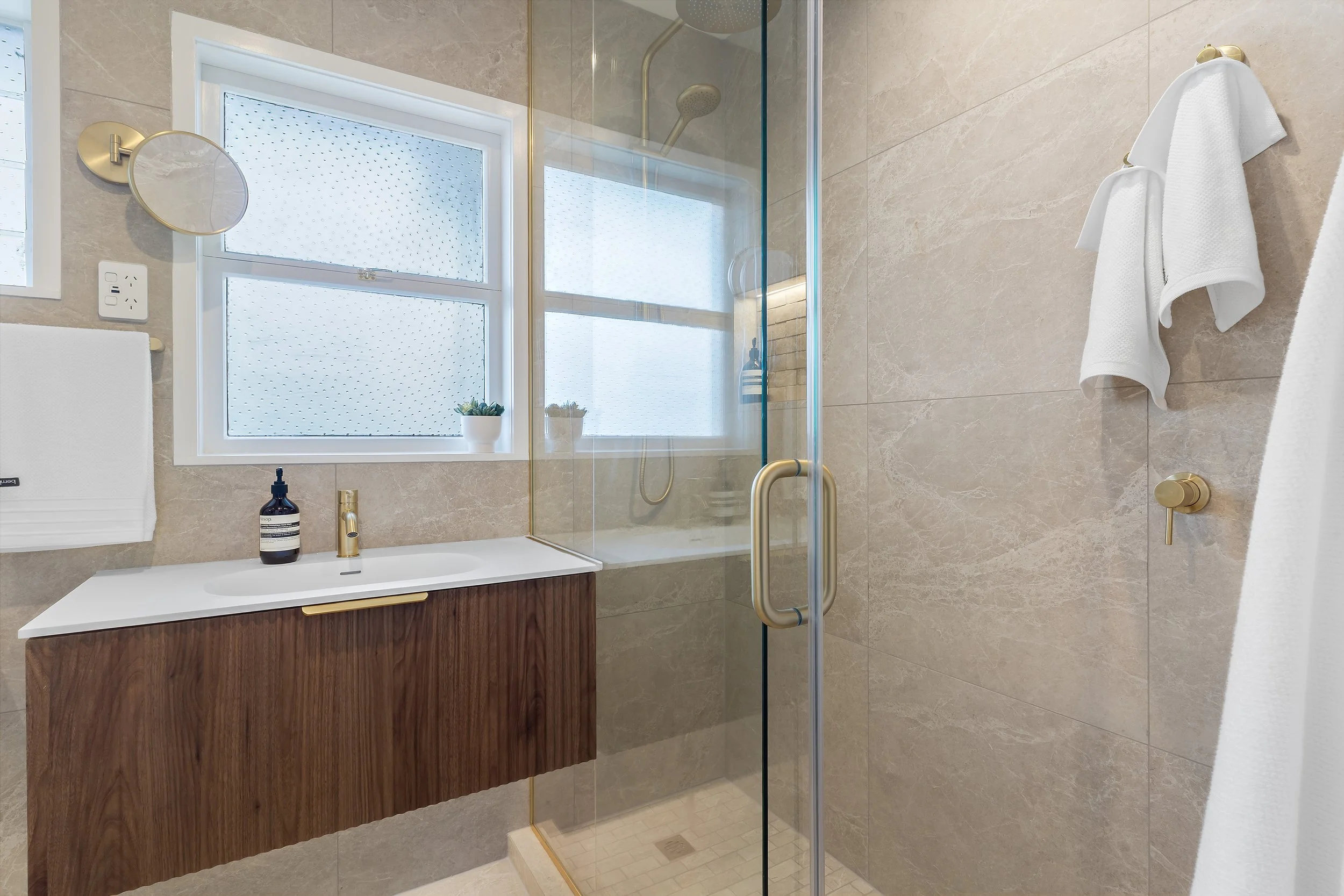 The simple wooden vanity and tower cabinet evoke a driftwood feel, seamlessly blending with the beige, sand-colored tiles and mosaics make you feel like you are stepping into a coastal retreat. This curated mix immediately delivers a calm, beach-insp