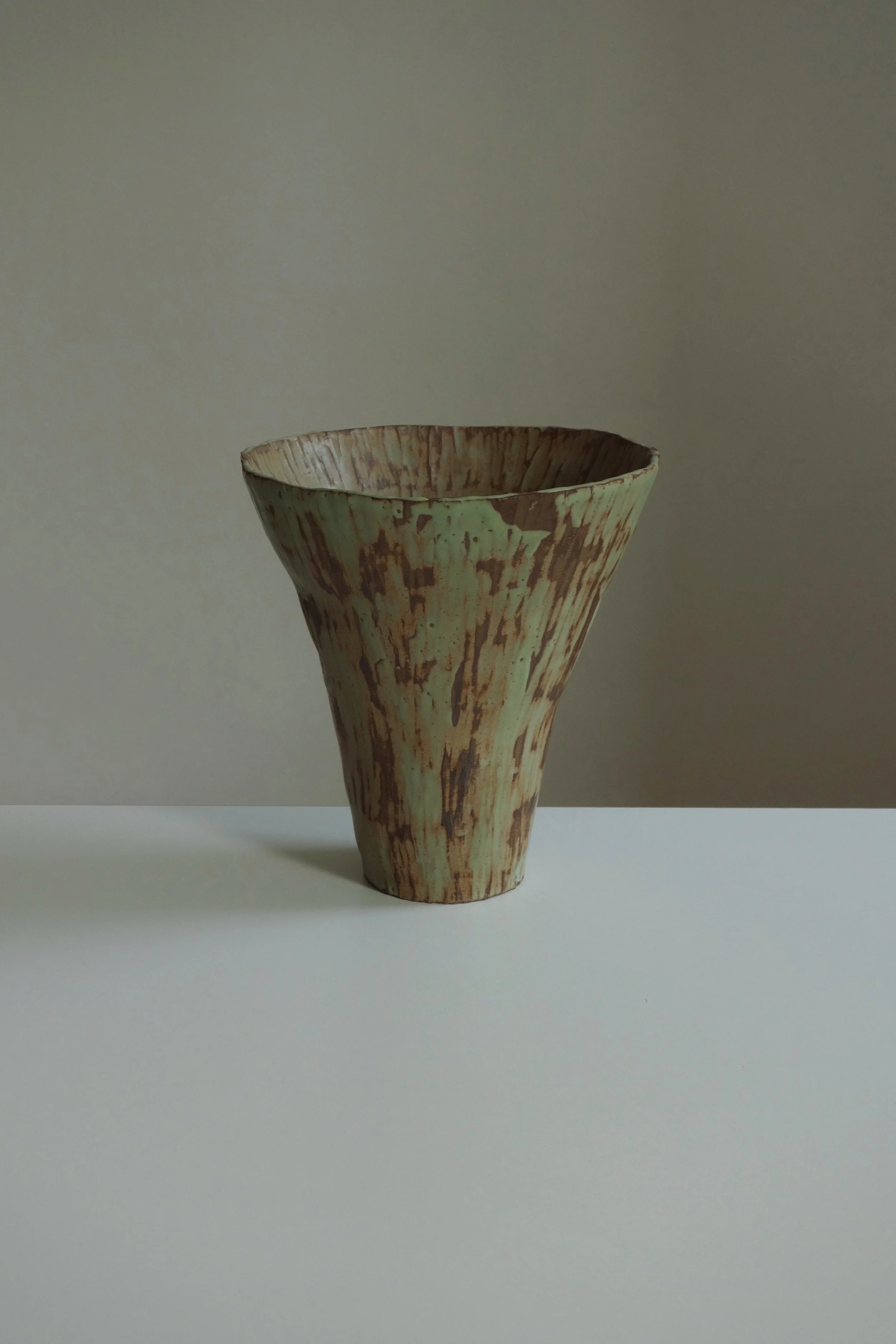 A ceramic vase with a textured, earthy design, placed on a white surface against a plain, light-colored background.