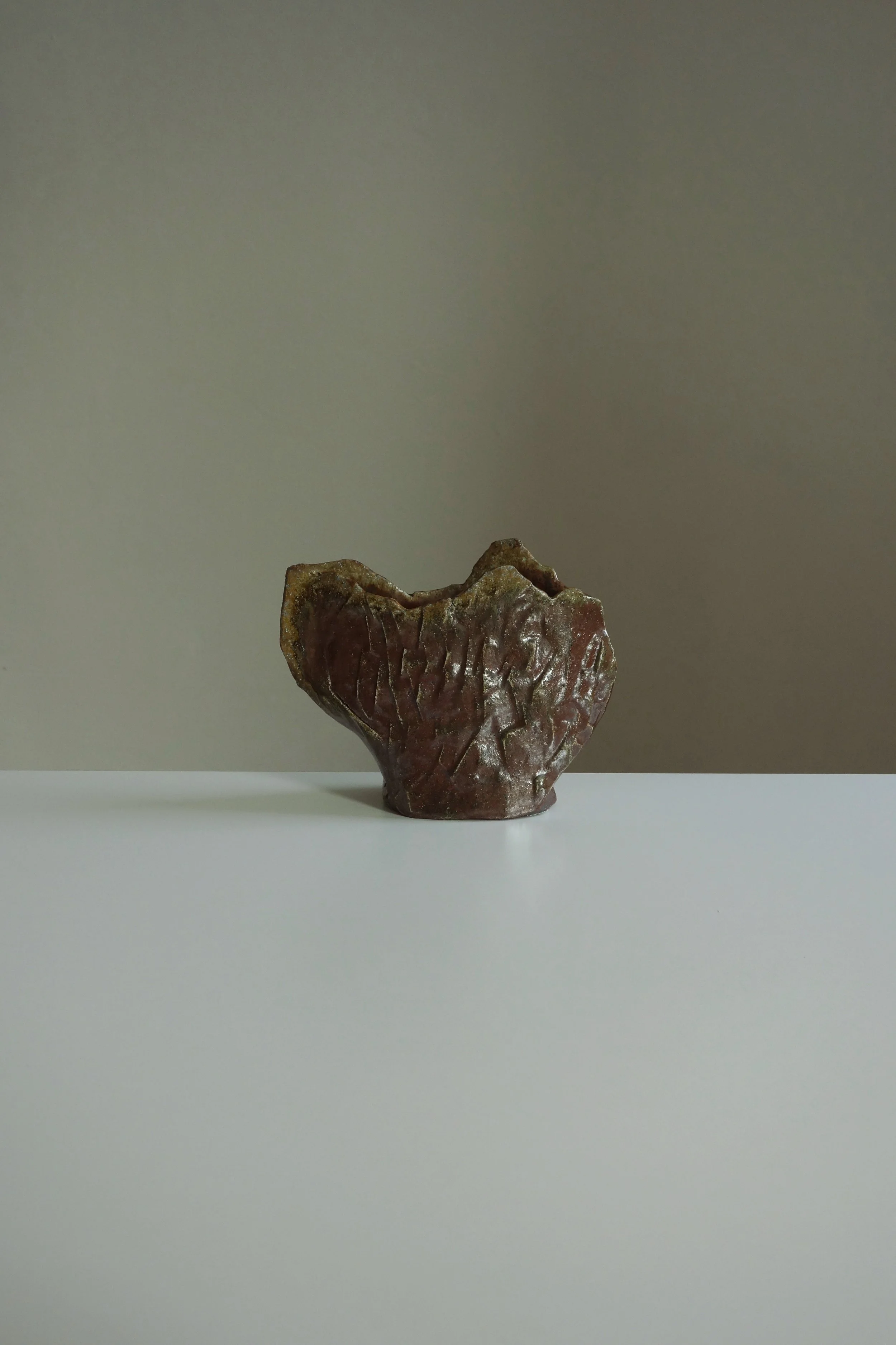 A small, irregularly shaped ceramic or clay vessel with a textured, reddish-brown surface, placed on a white surface against a neutral gray background.