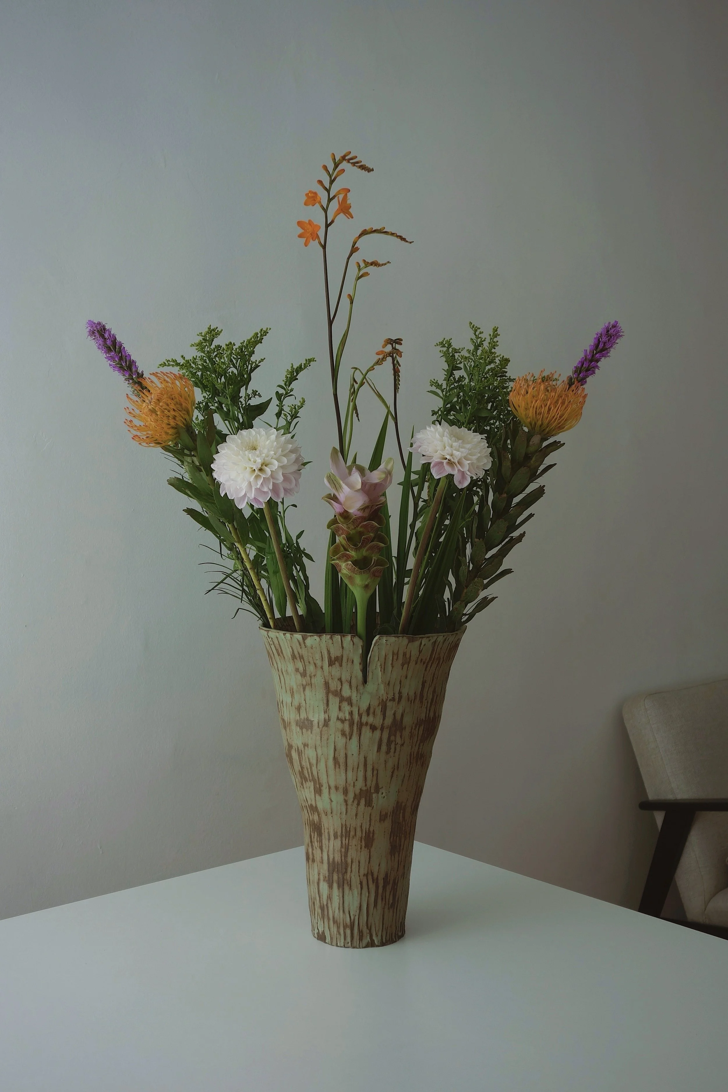 A tall beige vase with a wood grain pattern, filled with various colorful flowers including purple, orange, pink, and white blooms, placed on a white table against a plain light-colored wall.