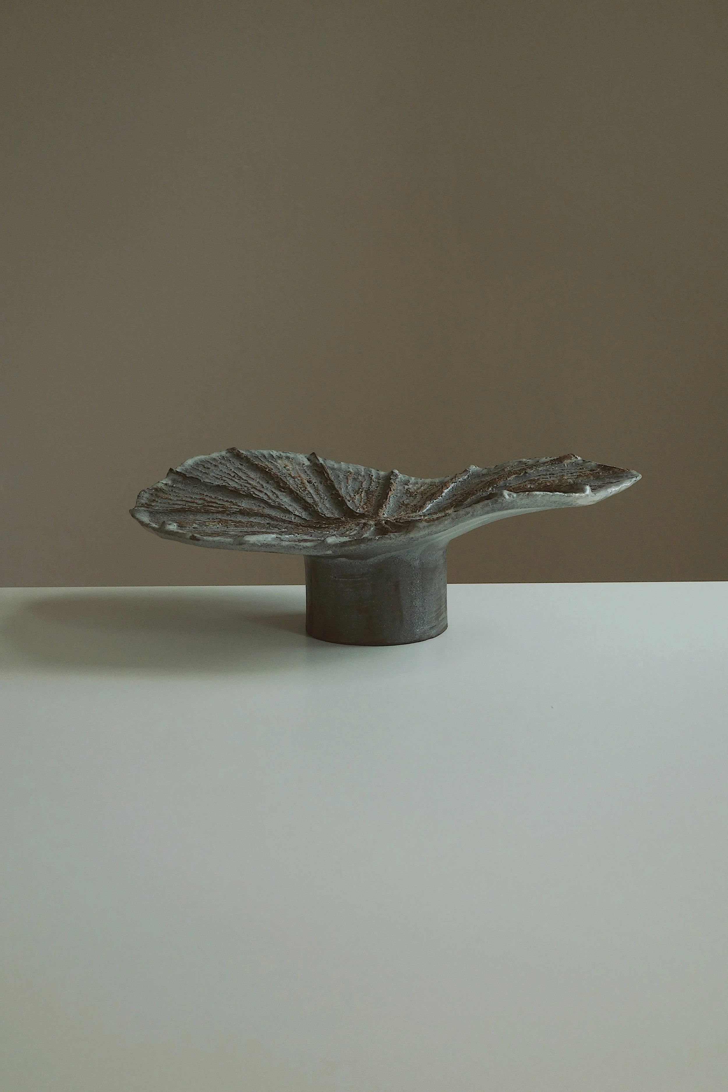 A ceramic sculpture resembling a leaf on a short cylindrical base, placed on a white surface with a beige background.