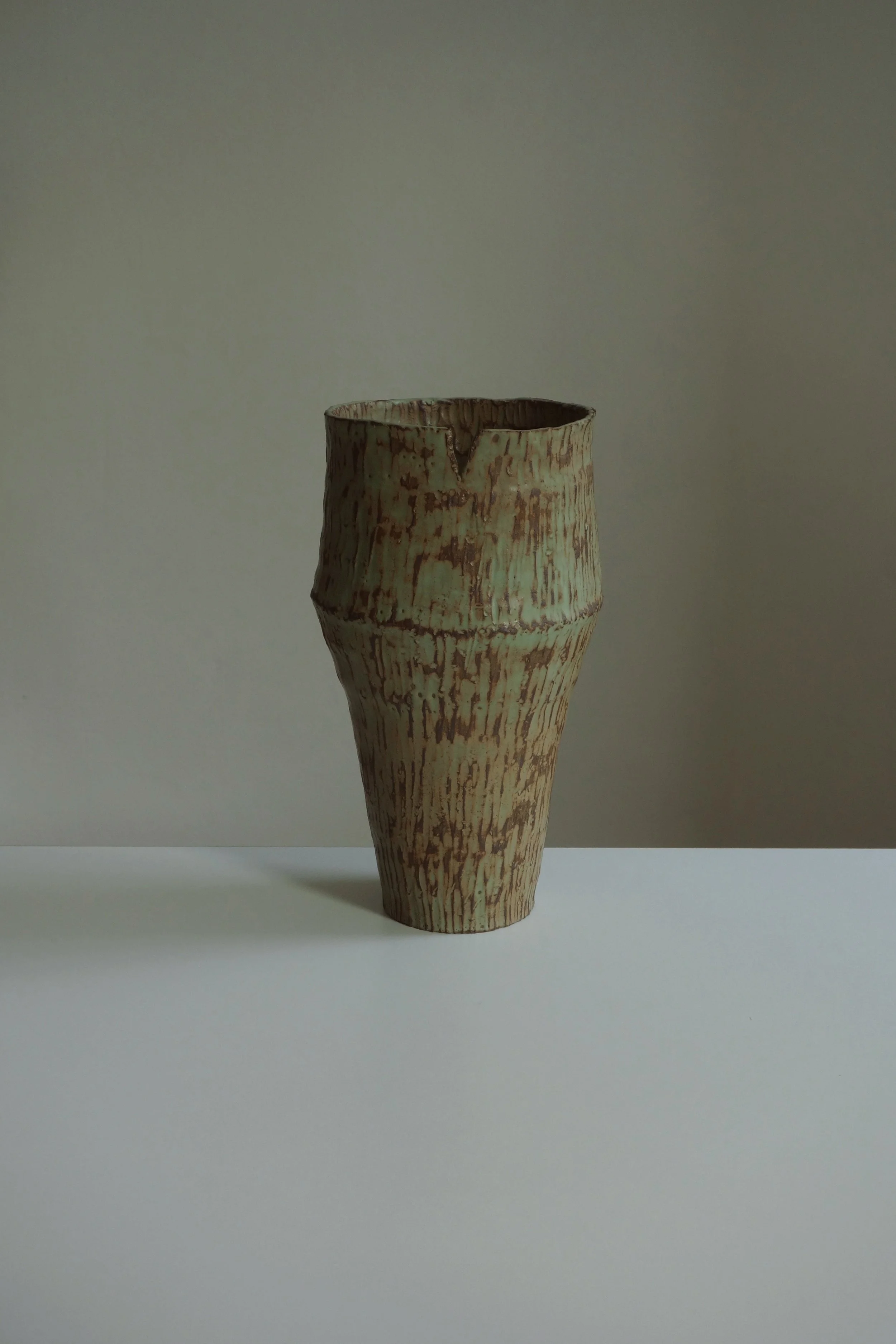 A tall ceramic vase with a textured, uneven surface and a beige and brown mottled glaze, placed on a white surface against a neutral background.