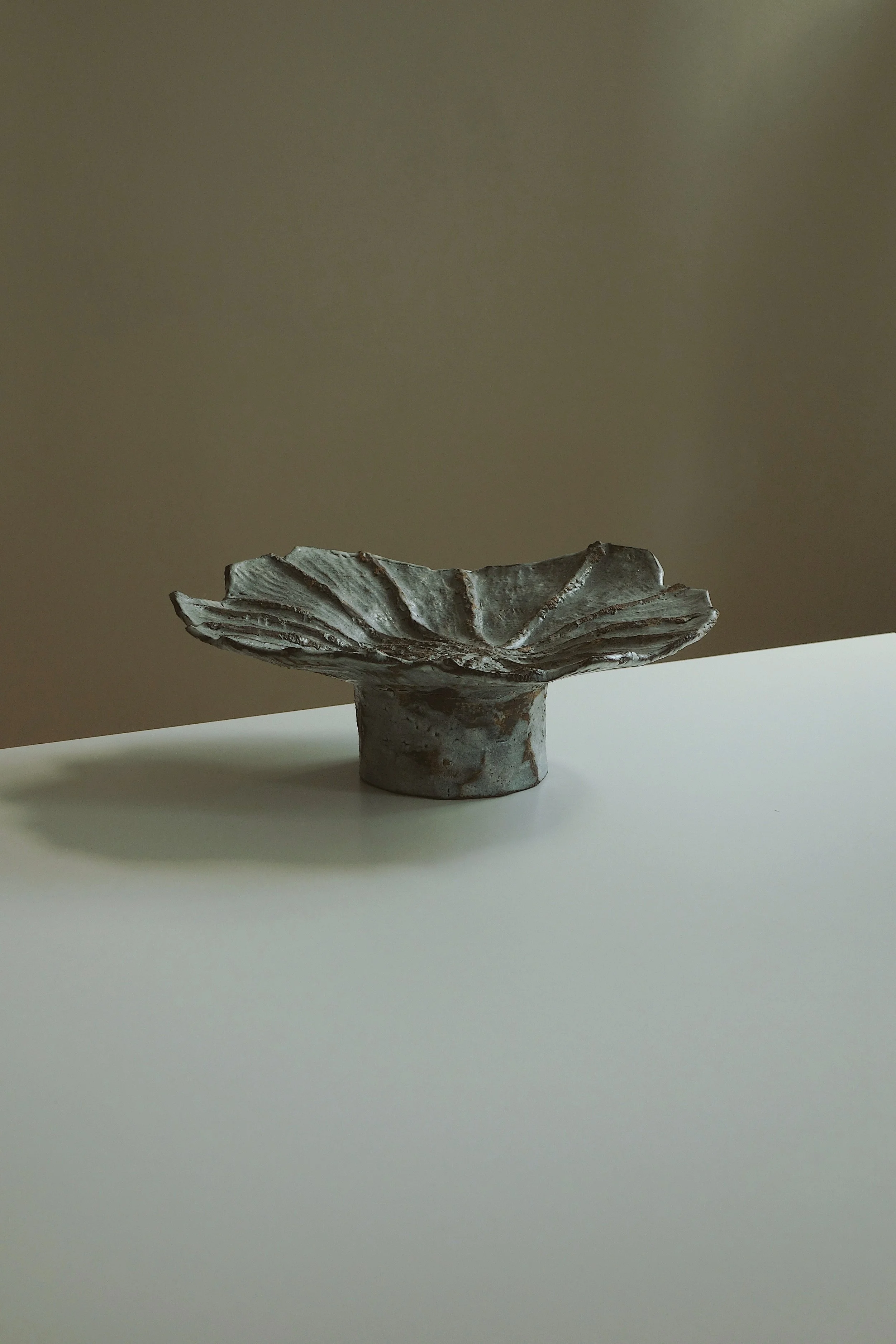 A ceramic sculptural bowl with an organic, leaf-like shape and textured surface, placed on a white surface with a plain background.