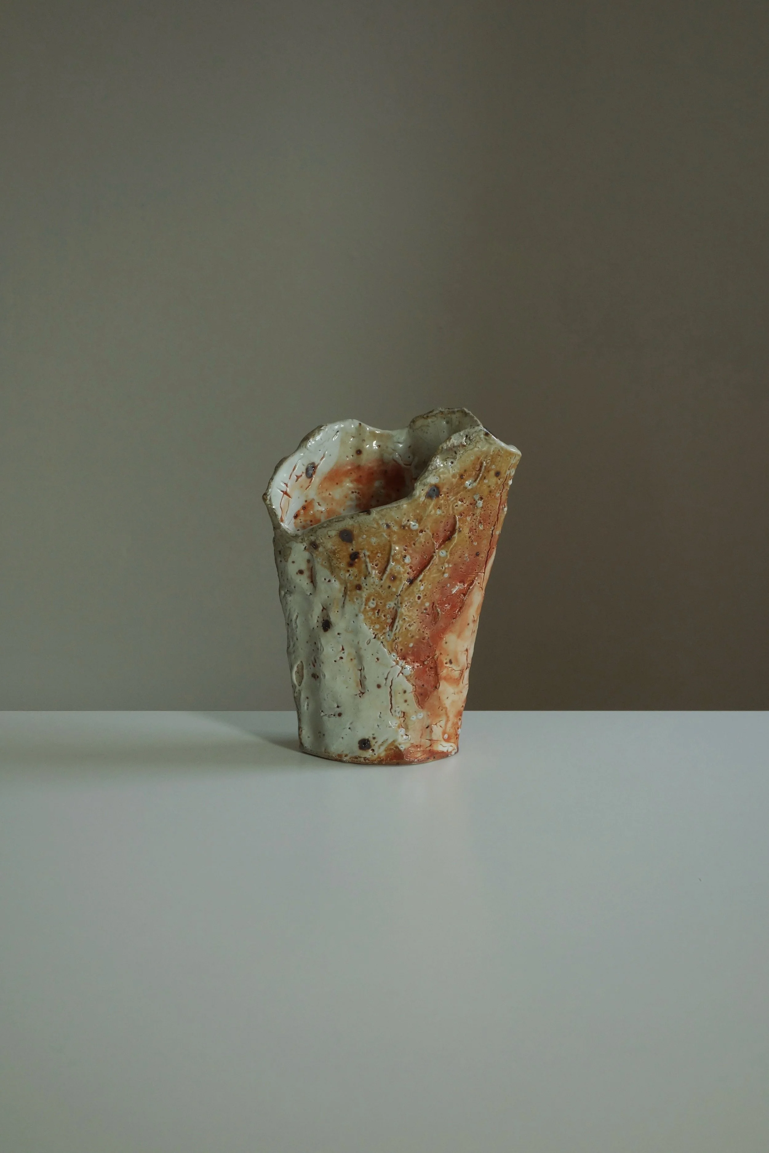 A ceramic sculpture with a textured, irregular surface, featuring beige, orange, and white colors, placed on a white table against a neutral background.