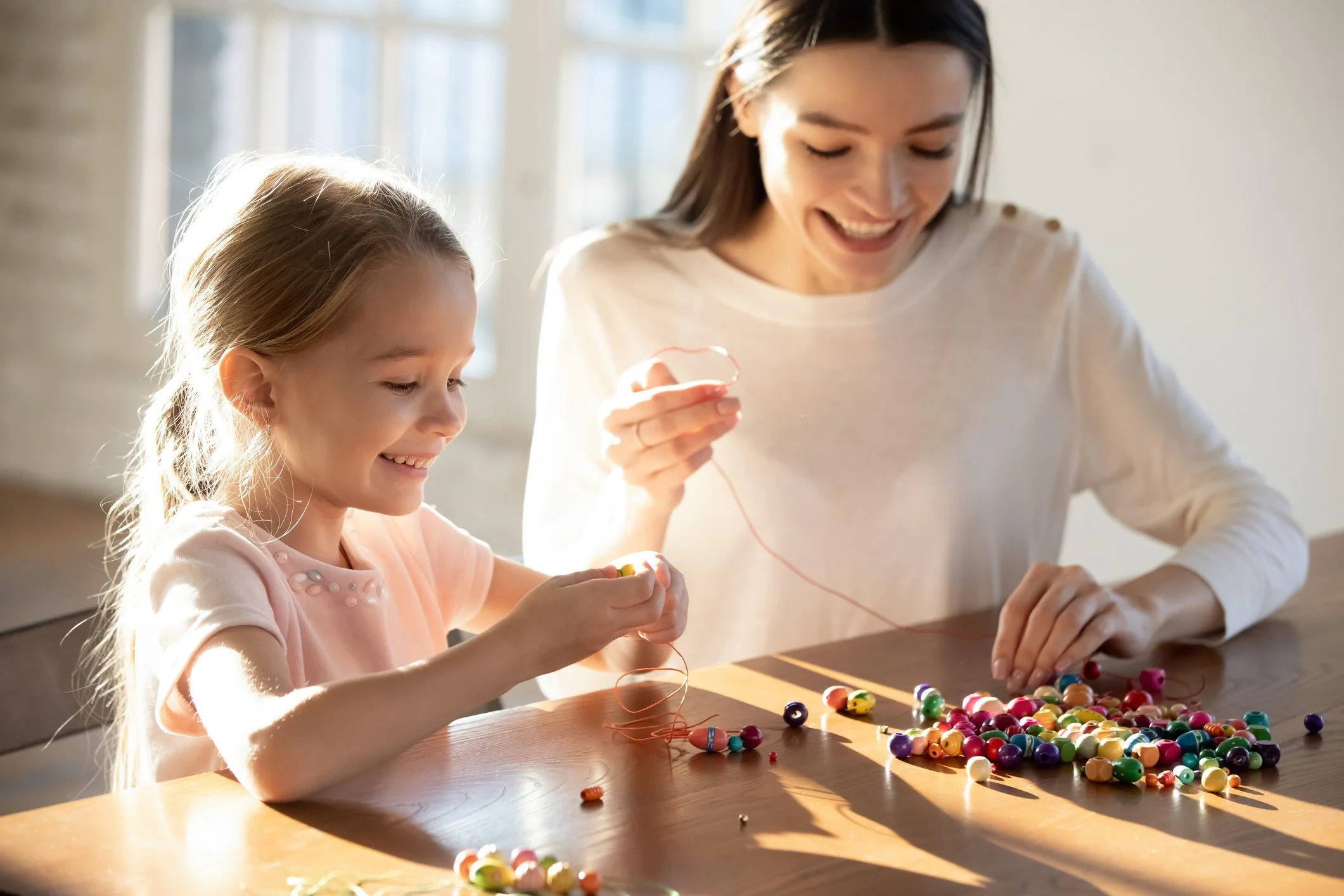 A young girl and woman playing with colorful beads at a wooden table in a sunlit room, smiling and enjoying a craft activity.