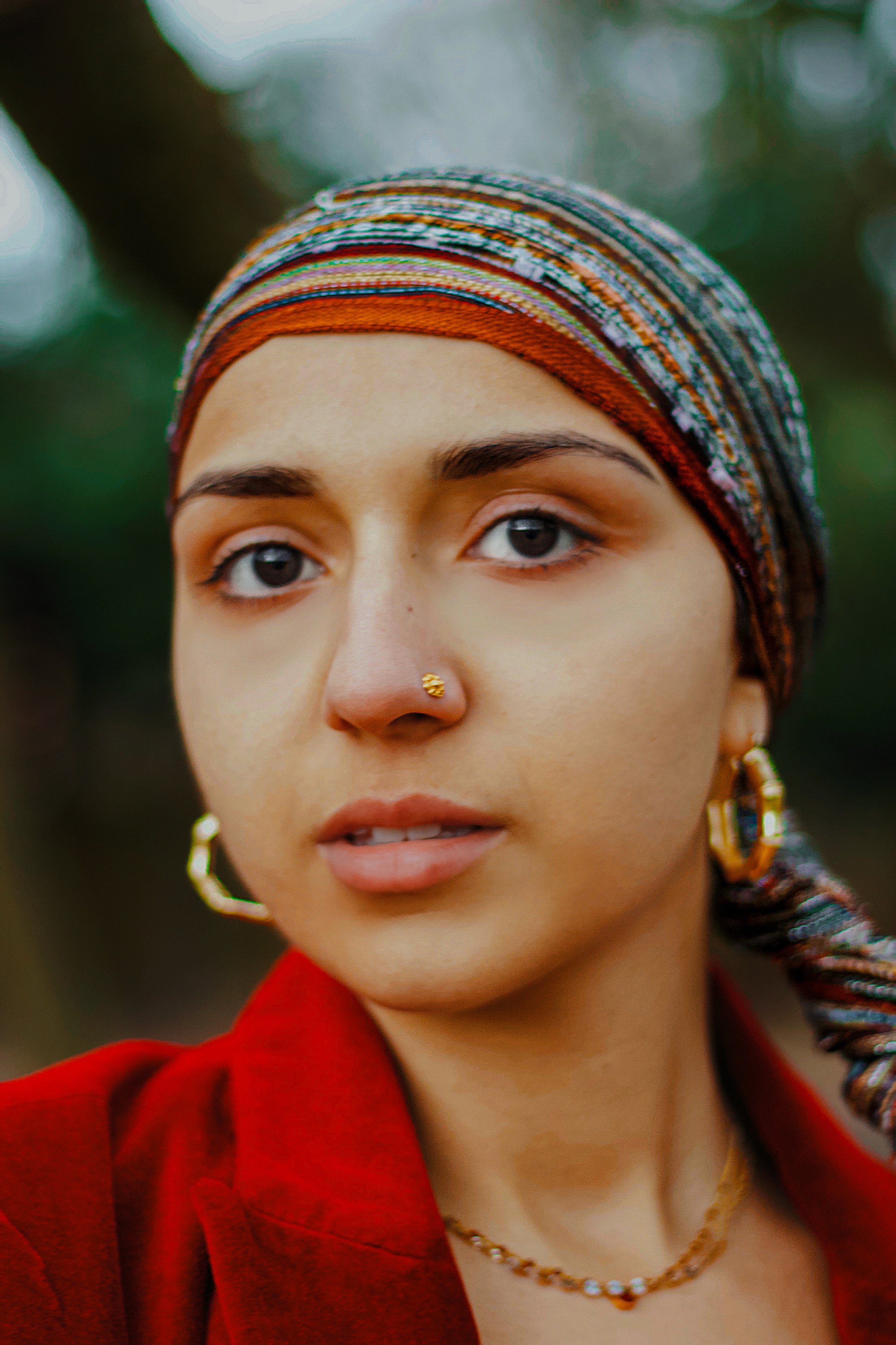 emotive headshot of indian woman  with a multicolored headscarf, gold jewelry, and a red blazer and gold indian jewellery (nose ring and hoops). Warm tones and calming curious expression