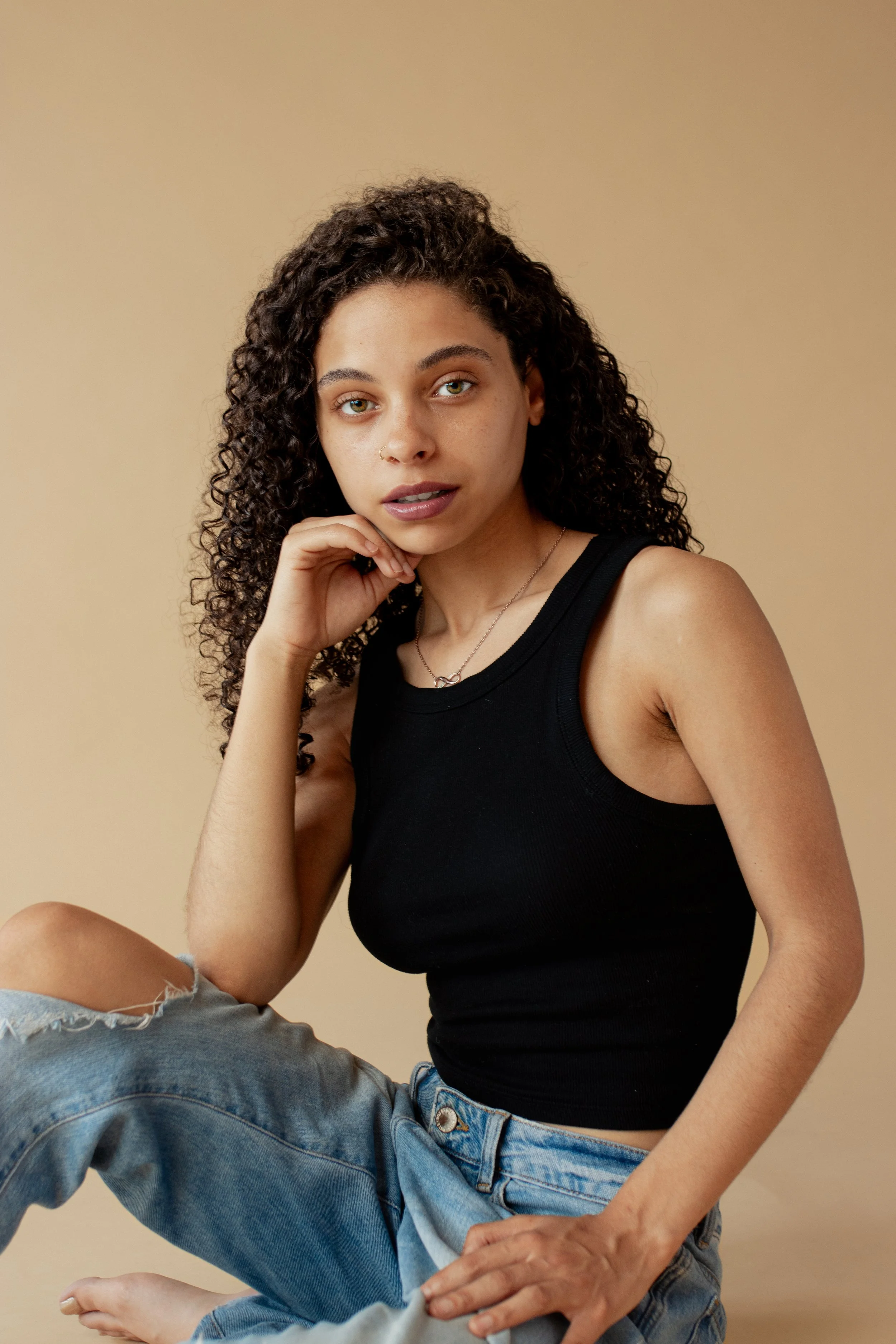 Kira Lei model, captured emotively with fresh vibrant lighting. A young woman with curly hair wearing a black tank top and ripped jeans, sitting against a beige background, looking at the camera with a relaxed expression.