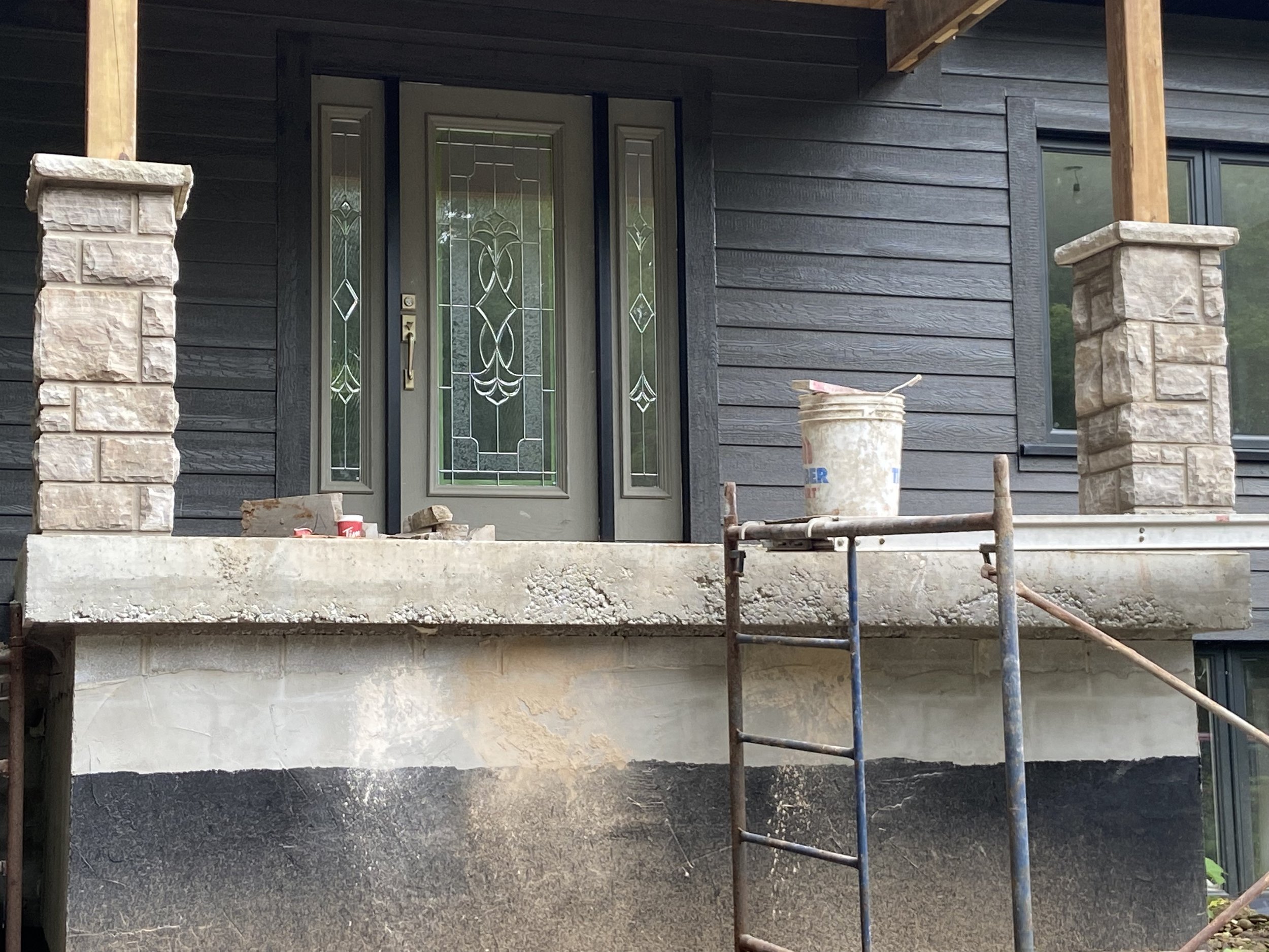 Front porch of a house under construction, featuring a decorative glass door, stone pillars, a concrete platform, and construction materials such as a bucket and scaffolding.