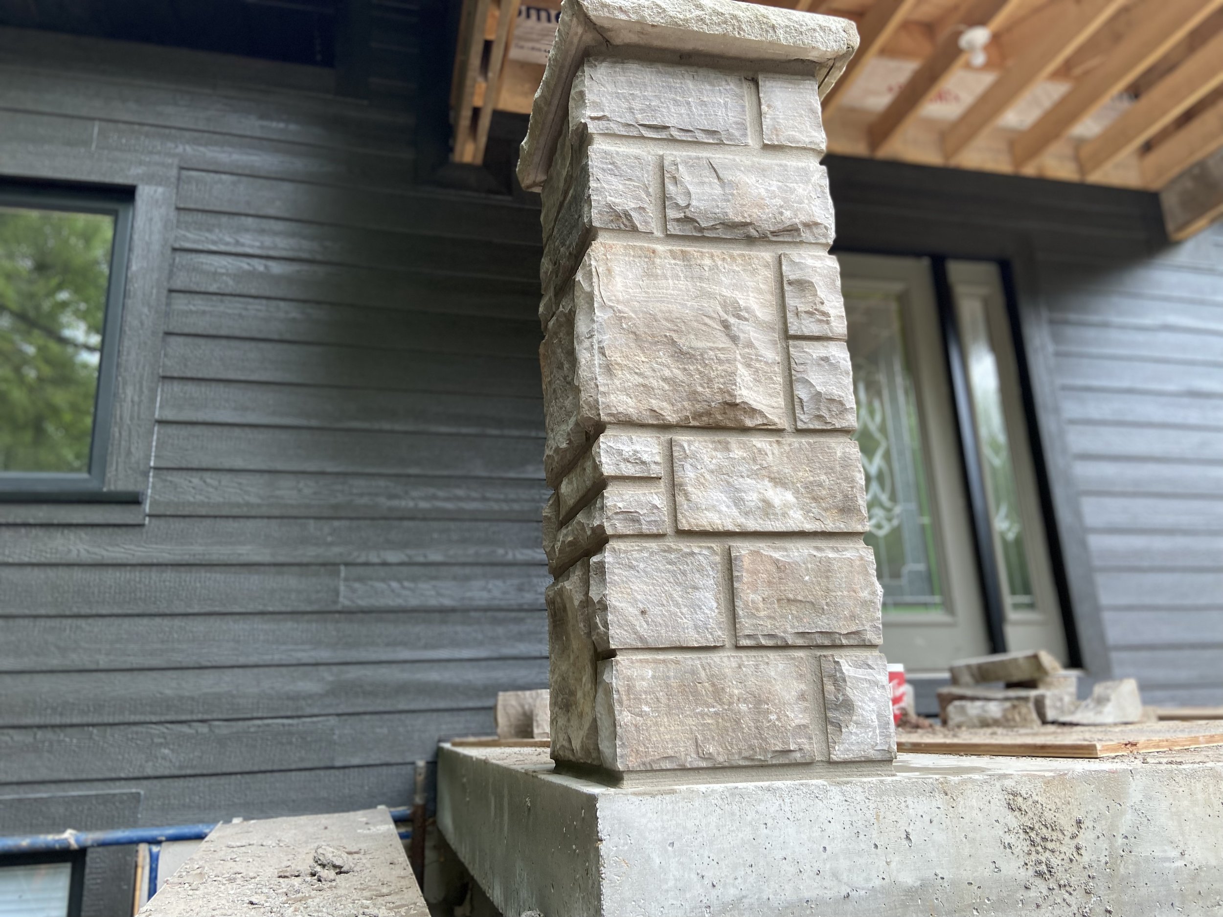 Stone pillar on porch under construction with gray siding and a window in the background.