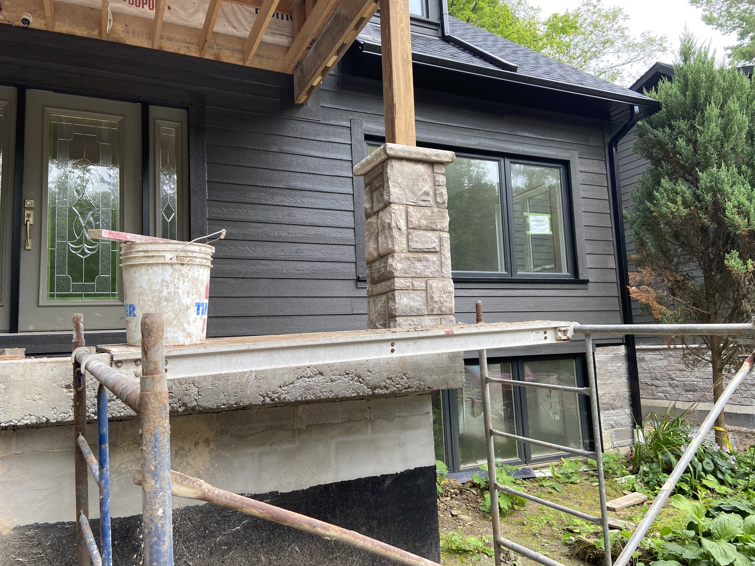 Construction scaffolding and a bucket in front of a house with dark siding and large windows.