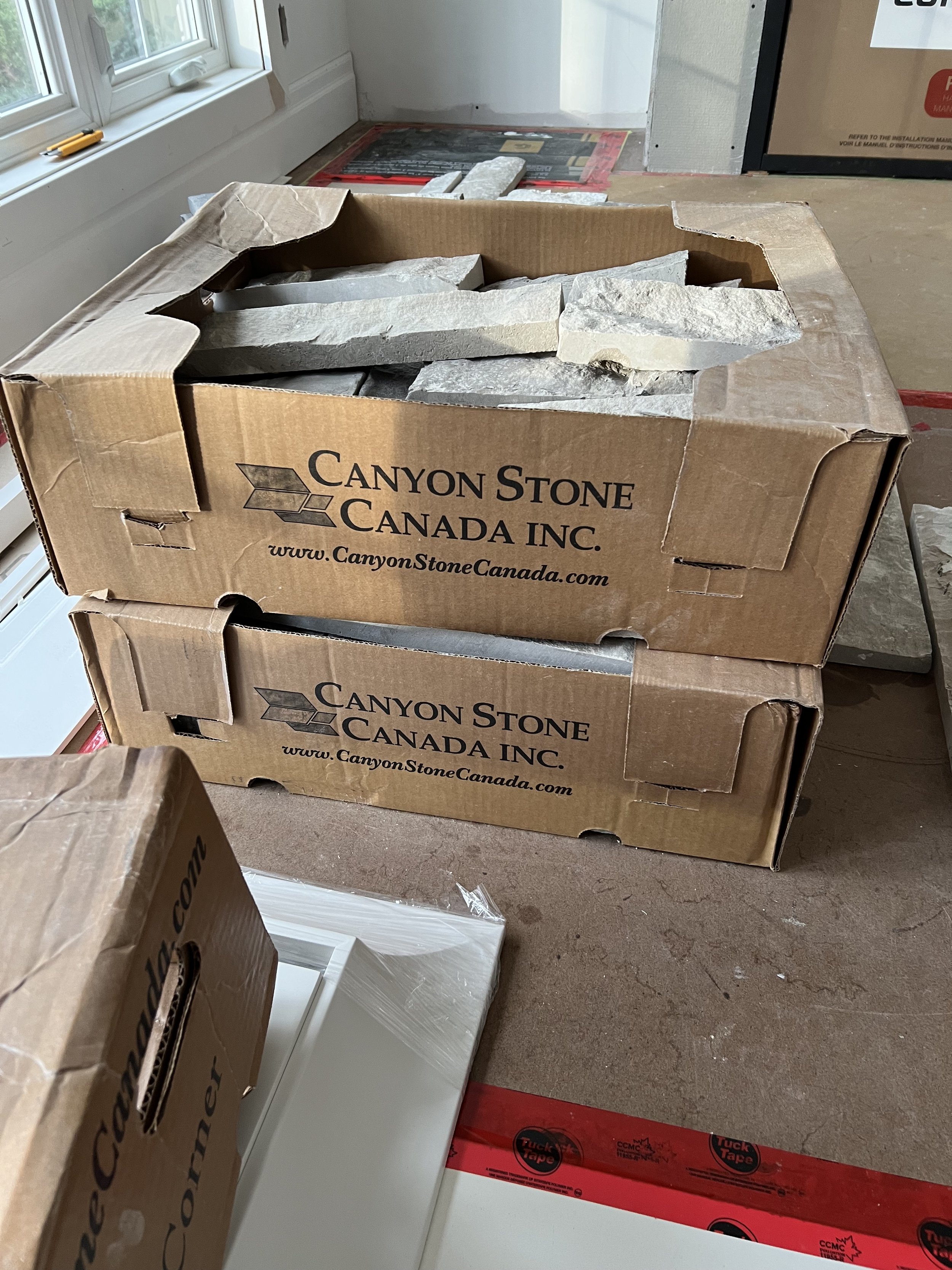 Cardboard boxes labeled "Canyon Stone Canada Inc." with stone pieces inside, sitting in a room under renovation.